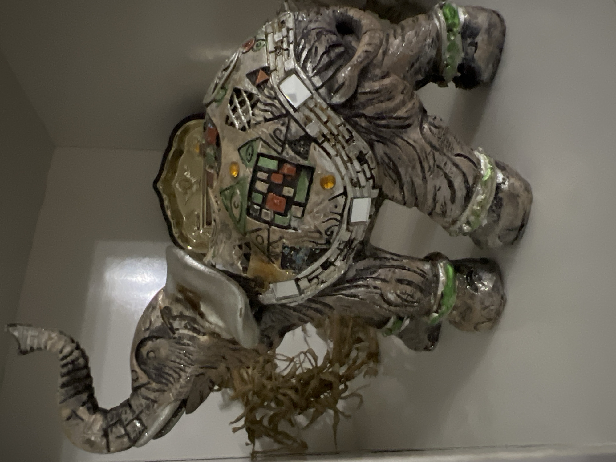 The image depicts a decorative elephant figurine with mosaic-style ornamentation on its body and a fringed material underneath. The figurine is gray with green detailing around its legs. It is positioned inside a white enclosed space, likely a shelf or display case. The location is not discernible from the image.