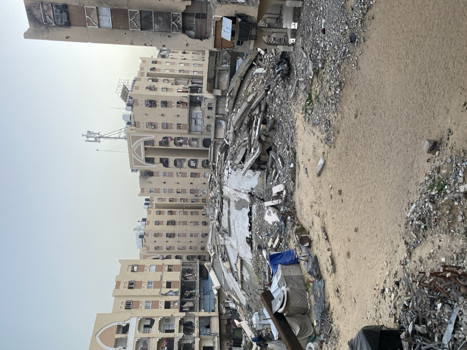 This image depicts a wide view of an extensively damaged urban area in Khan Yunis, Palestinian Territory. The scene is dominated by widespread destruction, with a large, central expanse of pulverized building materials, concrete slabs, and twisted rebar indicating the complete collapse of one or more structures. This central rubble field is surrounded by multiple multi-story residential buildings, primarily light tan in color, which stand partially damaged. These surviving structures exhibit numerous projectile impacts or shrapnel holes across their facades, blown-out windows, and exposed interiors on various floors. Some buildings show evidence of charring or fire damage, particularly on their upper levels. Rooftops are visible with water tanks, satellite dishes, and a prominent communications tower with multiple antennae. In the lower-left foreground, several makeshift shelters constructed from blue tarpaulins and other materials are present amidst piles of debris and salvaged items. The ground throughout the foreground is a mix of disturbed sand, dirt, and scattered rubble. No individuals are visible in the frame, and no specific actions or interactions are depicted. The sky is overcast or hazy, providing soft, diffused lighting.