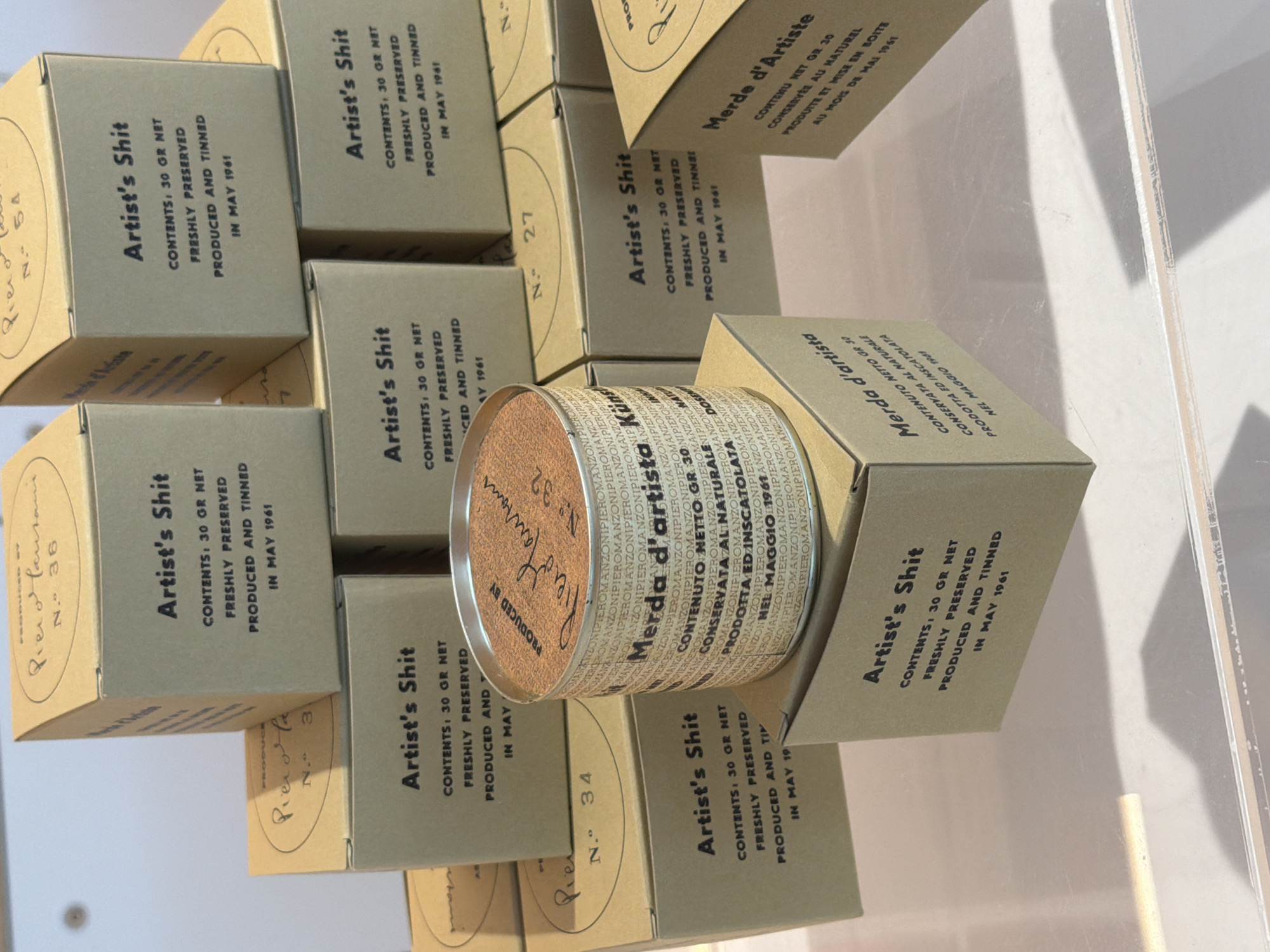 This image depicts a display of artist's materials, likely a curated exhibition or shop display, in Cold Spring, United States. The primary subjects are numerous small cardboard boxes and one cylindrical tin, all featuring similar text printed in black. The text on many of the boxes reads "Artist's Shit," followed by details about contents, preservation, and production date, often "IN MAY 1961." Some boxes also have handwritten text and a number, such as "No. 36," "No. 34," and "No. 27." The cylindrical tin, centrally located and opened, displays similar text in Italian, "Merda d'artista," and contains a reddish-brown powdery substance. The boxes are stacked in a layered arrangement, creating a sense of depth. The overall color palette is dominated by the earthy tones of the cardboard and the black text. The lighting suggests an indoor environment, possibly a gallery or retail space, with a light-colored surface or display table and some soft shadows. The background is out of focus, indicating the focus is on the displayed items. There are no people visible in the image.