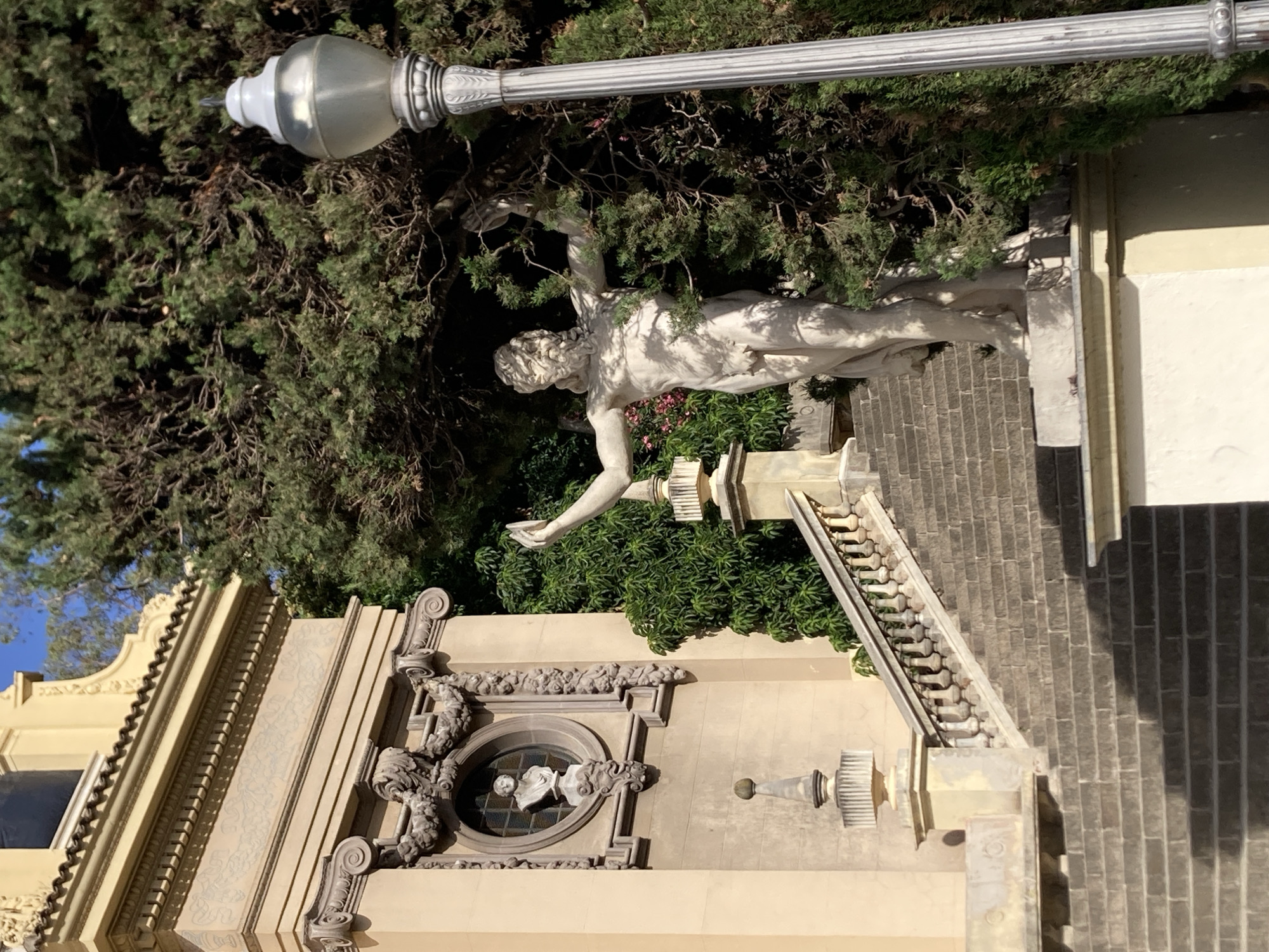 The image shows a marble statue of a muscular, bearded man with outstretched arms, partially obscured by lush green trees. The statue stands on a stone pedestal on the left side of a wide stone staircase. To the left of the staircase is a light-colored building with ornate architectural details, including columns, cornices, and a circular window containing a bust. A modern street lamp with a silver finish stands on the right side of the frame, positioned in front of the trees. The sky is visible at the top of the image, indicating an outdoor setting. This is likely Montjuïc Hill in Barcelona, Spain, given the architectural style and the presence of such statues and gardens.