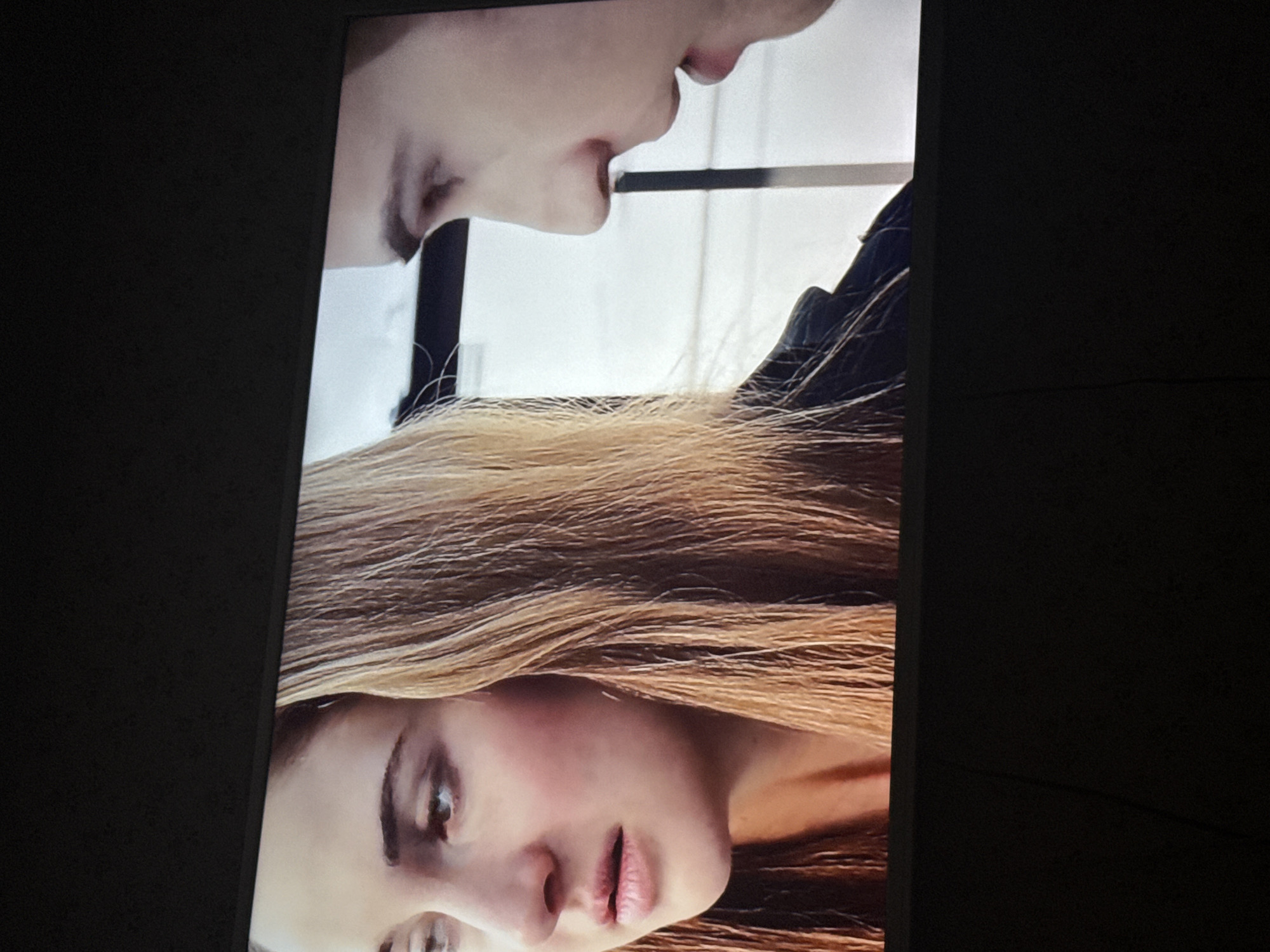 A still image, possibly a screencap from a movie or television show, depicts a close-up of two individuals. A fair-skinned woman with long, blonde hair is positioned at the bottom, facing the camera with a downward gaze. Above her, and partially overlapping her, is another fair-skinned individual whose face is partially visible, focusing on their mouth and jawline. The background is a mix of white and dark, vertical lines. It is impossible to discern the location within Mytishchi, Russia based on this visual data.