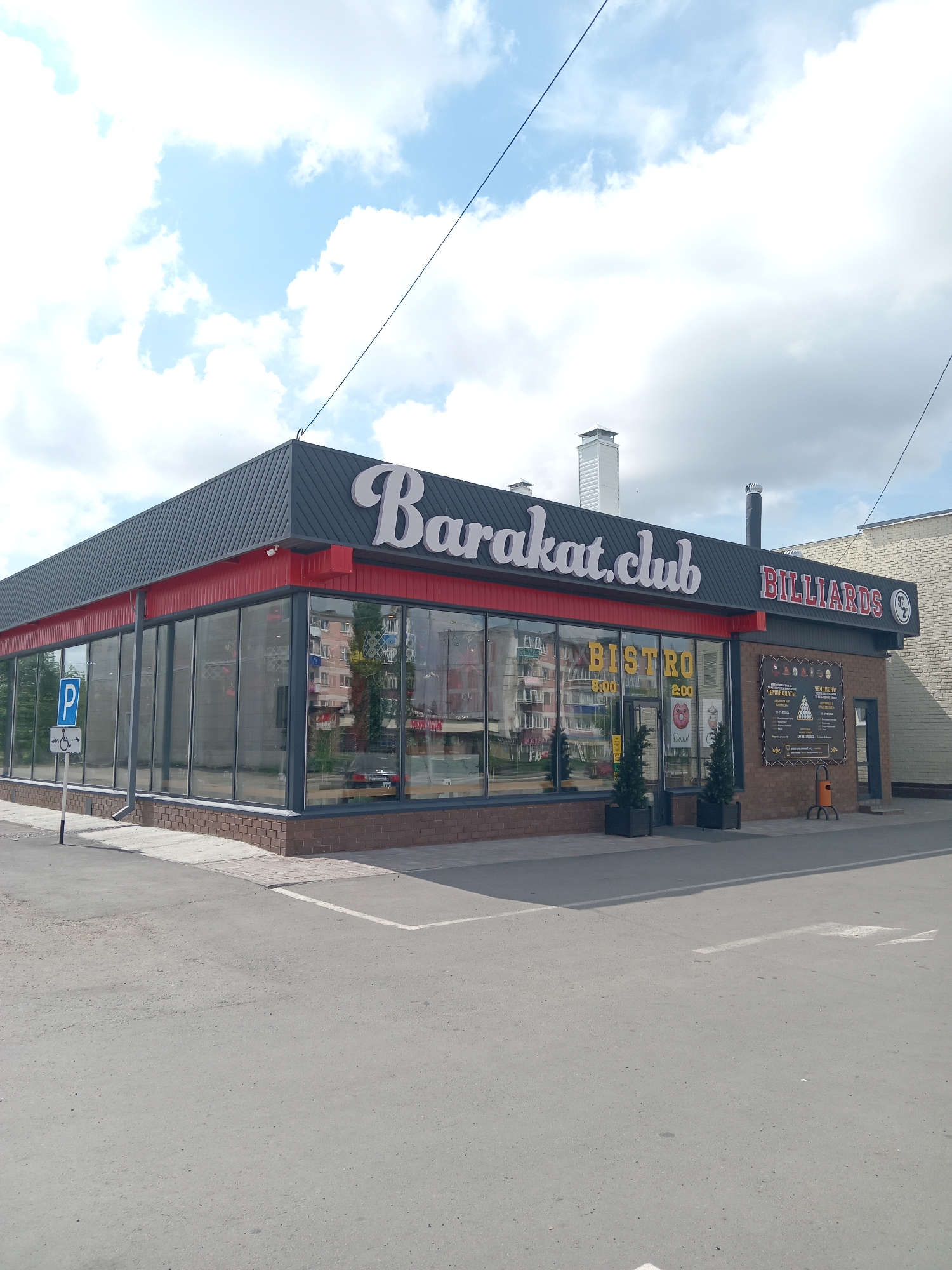 A daylight, eye-level shot shows the exterior of a commercial establishment named "Barakat.club Billiards" in Rudny, Kazakhstan. The building features a modern design with large glass windows and a dark grey, sloped roof. Red trim accents the building's facade. Prominently displayed on the roof are the words "Barakat.club" in large, white, stylized font and "BILLIARDS" in red, block letters.
To the left of the entrance, a blue sign with a white 'P' indicates parking, accompanied by a sign denoting accessibility for individuals with disabilities. Two dark green, potted evergreen trees stand in front of the entrance.
Visible through the glass windows are interior elements, including a "BISTRO" sign with operating hours, and what appears to be billiard tables. On the brick wall to the right of the entrance, a poster advertises a billiard championship. An orange trash receptacle is situated near the entrance.
The environment includes an asphalt parking lot in the foreground and a bright blue sky with scattered white clouds. Residential buildings are reflected in the glass facade. The overall scene suggests a commercial area in Rudny.