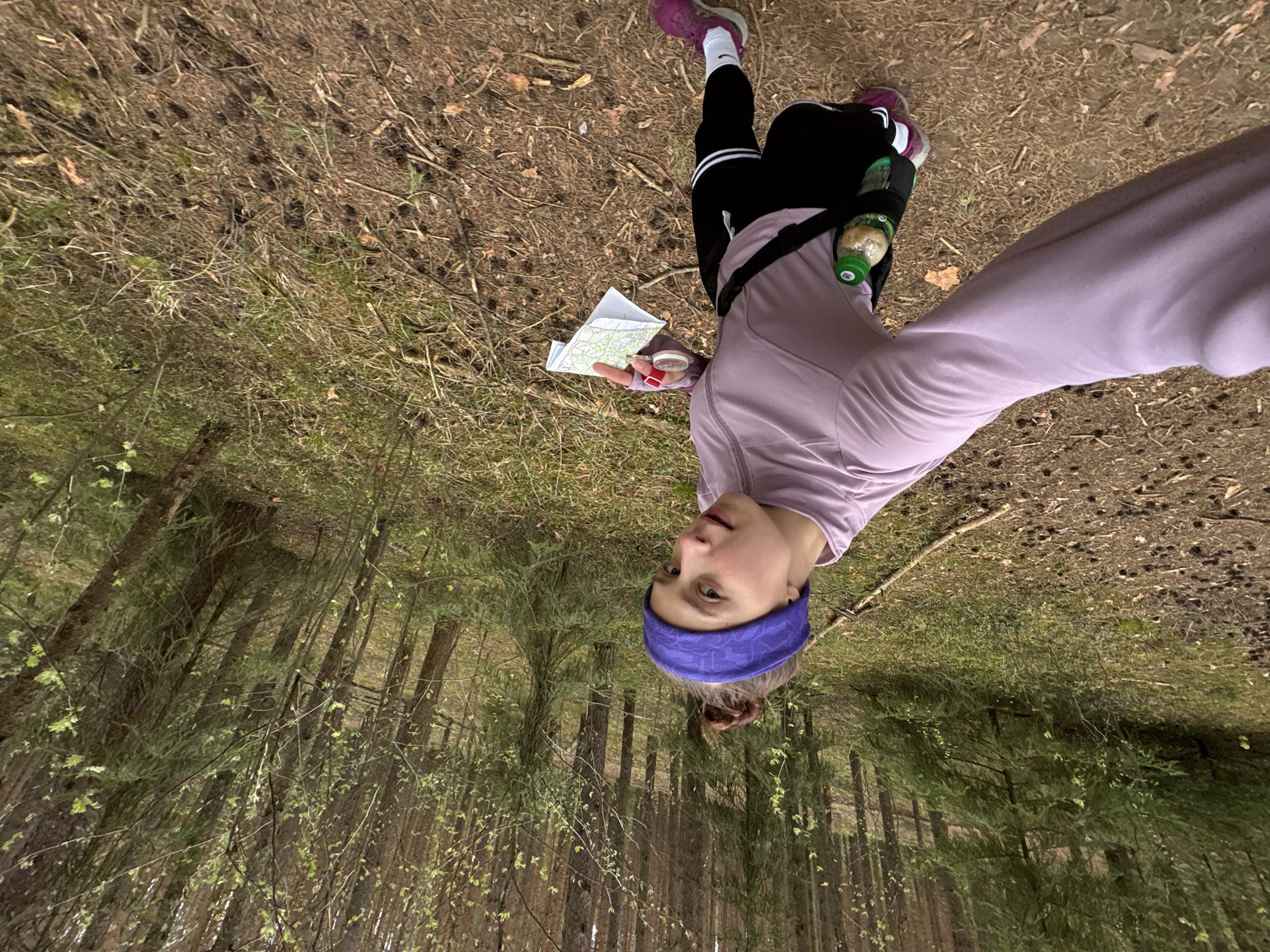 A young woman stands in a pine forest, possibly a city park. She is wearing a purple athletic headband, a long-sleeved lavender shirt, black athletic pants with white stripes, and pink running shoes. She holds a folded map and a compass in her left hand. A water bottle is secured to her body by a strap across her chest. The ground is covered with pine needles and sparse vegetation. The trees are tall and slender, typical of a pine forest. The scene appears to be captured with a selfie perspective. The location is unspecified beyond Russia, and lacking specific details no precise location within Perm is identifiable.