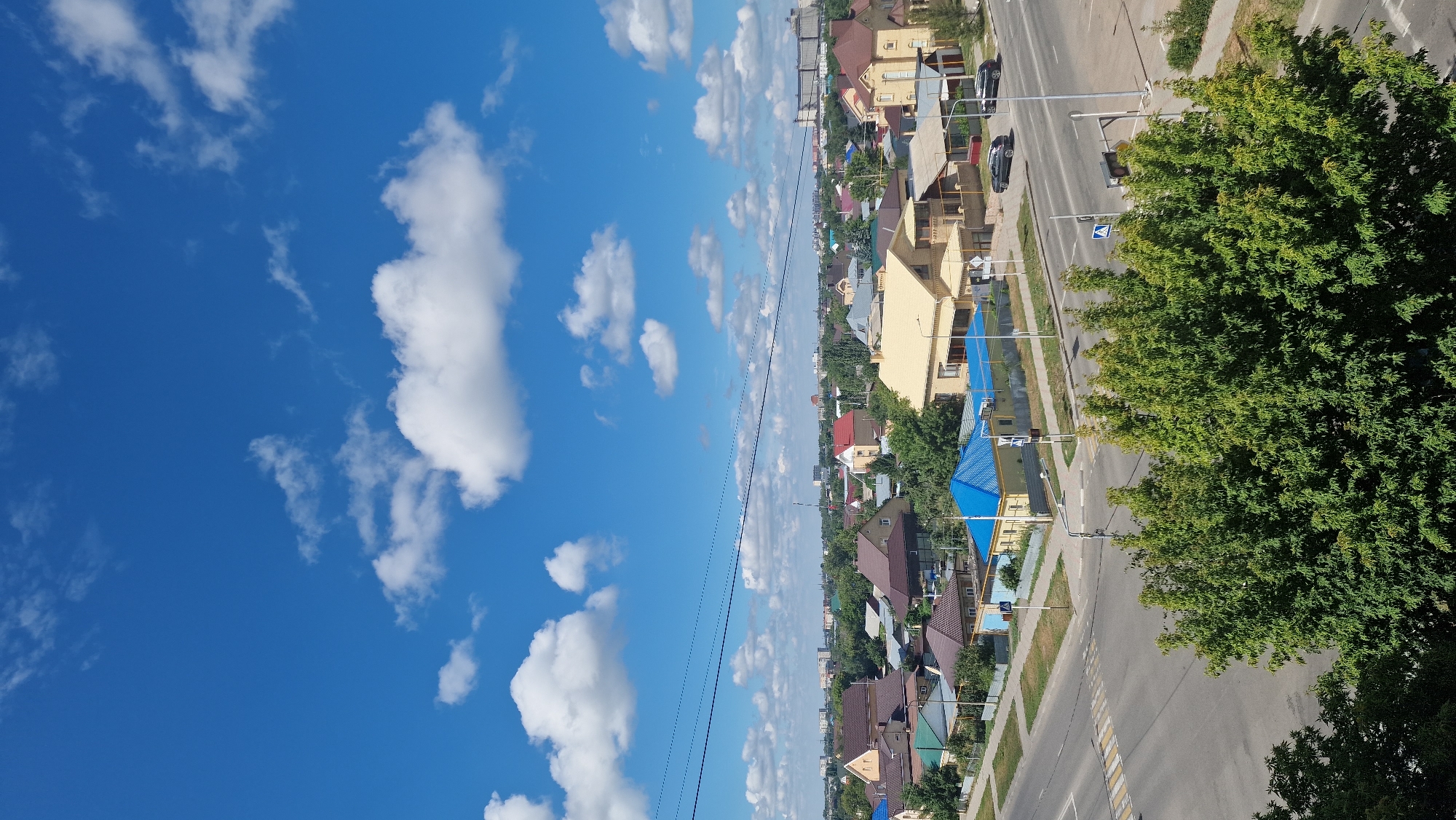 The image depicts a suburban landscape under a clear blue sky with scattered white clouds. In the foreground, a dense canopy of green trees partially obstructs the view of a road intersection. The intersection features a crosswalk and traffic signals. Beyond the road, a collection of residential houses with various roof colors, including brown, blue, and yellow, are visible. The houses are of different architectural styles and sizes, suggesting a mix of older and newer constructions. In the background, a more distant view of the city of Kostanay, Kazakhstan, can be seen, with apartment buildings and industrial structures forming the skyline. Two cars are parked on the side of the road. There are no people visible in the frame. The scene appears to be a typical sunny day in a residential area.