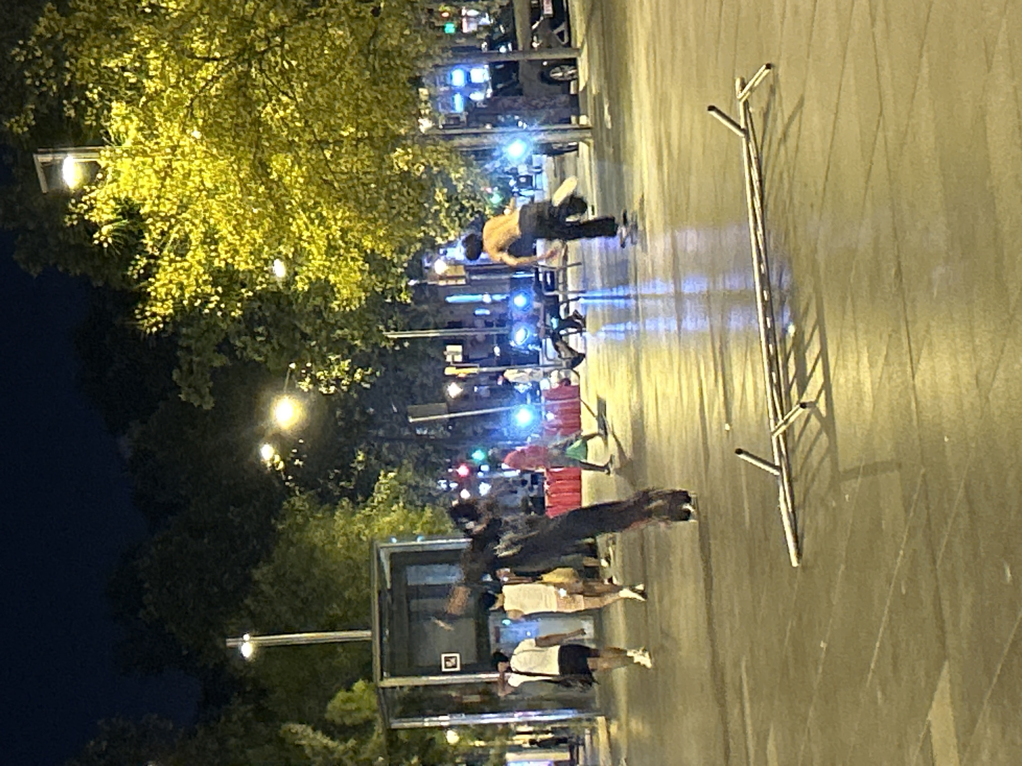 This image shows a nighttime street scene in Barcelona, Spain. In the foreground, a metal barricade lies on the ground. Further back, a person on a skateboard is performing a trick. Several other individuals are walking in the background. The scene is illuminated by streetlights and lights from buildings, casting long shadows on the wet pavement. The environment appears to be an urban plaza or pedestrian walkway, with trees and building facades visible. Traffic lights and cars are also present, suggesting a public thoroughfare.