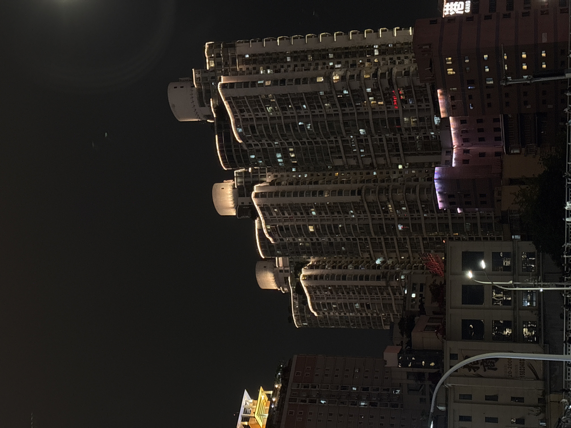 The image shows a night view of a city, identified as the Yuzhong District in China. The focus is on a set of high-rise residential buildings with distinctive, curved exterior designs accentuated by warm, ambient lighting along the edges. Adjacent to these buildings is a darker, more traditional-looking high-rise structure with lit windows and some signage at the top. Streetlights are visible at the lower right, casting a glow on the surrounding lower buildings and structures. The sky is dark, with what appears to be the moon or a bright light source at the upper left. The buildings are densely packed, showcasing a highly urbanized environment.