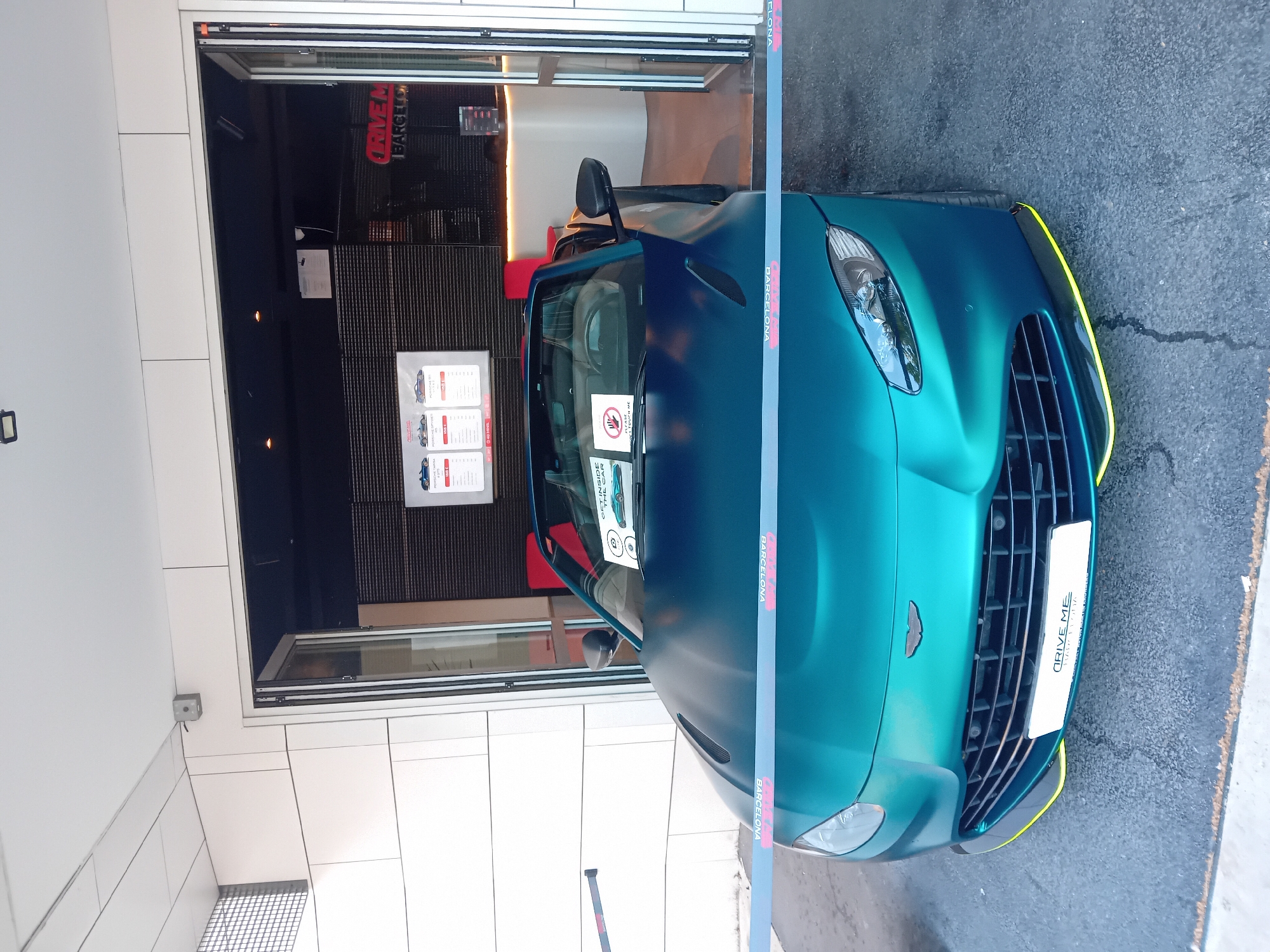 A vibrant blue Aston Martin convertible is parked in front of a business establishment in Barcelona, Spain. The car's license plate bears the words "DRIVE ME." A security ribbon printed with "BARCELONA" is stretched across the front of the vehicle, perhaps preventing access. The business, located behind the car, appears to be related to driving experiences, as evidenced by the signage "DRIVEM BARCELONA" and a price list featuring car images posted in the window. The surrounding environment is a modern building with a tiled façade and gray concrete pavement.