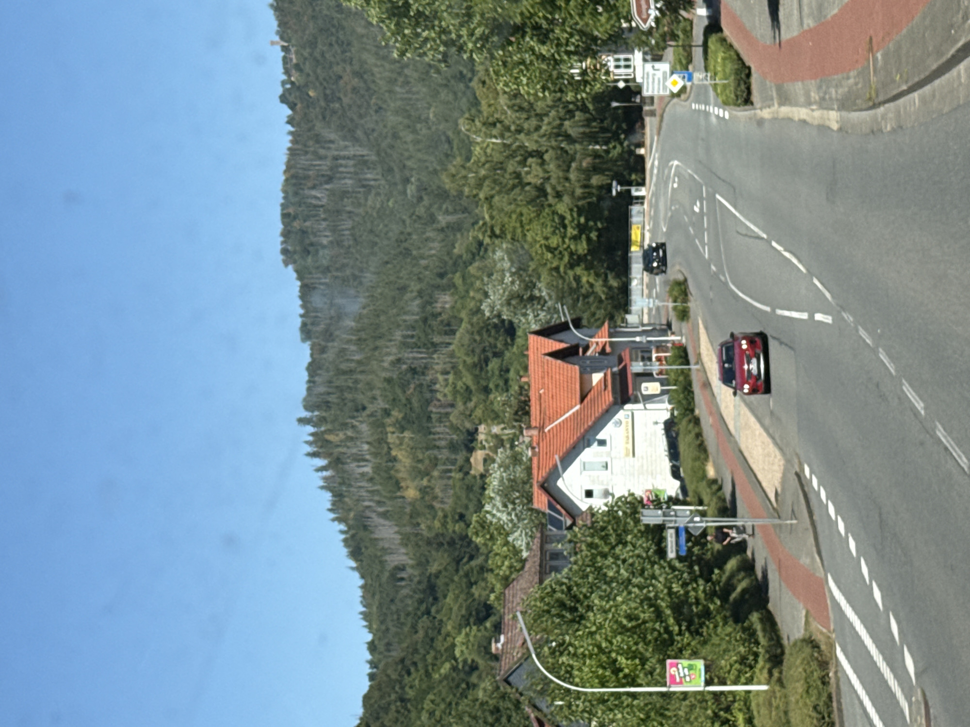 The image depicts a sunny day in Bad Lauterberg im Harz, Germany, with a road curving through a residential area at the base of a forested mountain. In the foreground, a paved road with white dashed lines runs from the lower left to the upper right of the frame. A red car is seen driving away from the viewer on the left lane. Further up the road, a black car is visible in the distance.
On the left side of the road, a sidewalk with a red-striped pedestrian crossing is present, bordered by lush green bushes. A white street lamp stands tall, adorned with a colorful, abstract sign. Adjacent to the sidewalk are houses with red-tiled roofs and white walls. One house has a sign with text on its exterior.
The background is dominated by a dense, green forest covering a steep mountainside. The mountain exhibits a sparse section of dead, grey trees, indicating a possible past event like a forest fire or disease. A tall, slender tower is visible on the very top of the mountain, far in the distance. The sky is a clear, bright blue. A person is seated on a bench on the left side of the road, near the street lamp, observing the scene.