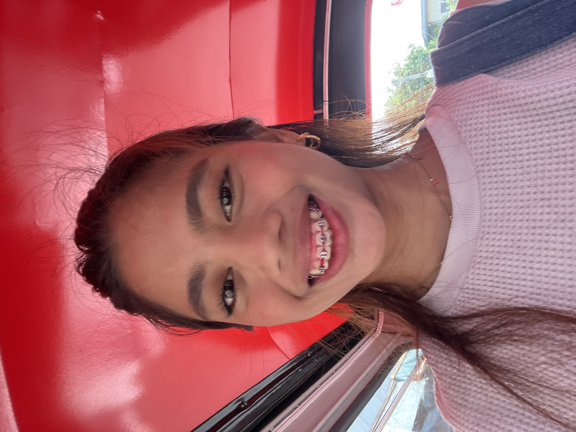 A young woman is taking a selfie in what appears to be a tuk-tuk, a motorized rickshaw, in Panabo City, Philippines. She has long brown hair, wears braces with pink bands, and a light-colored waffle-knit shirt. A gold hoop earring is visible in her left ear, and a delicate necklace with small orange charms hangs around her neck. She is looking directly at the camera with a wide smile. The interior of the tuk-tuk is bright red, and the background shows a glimpse of the outdoors, with greenery and buildings suggesting an urban setting. The lighting indicates it is daytime.