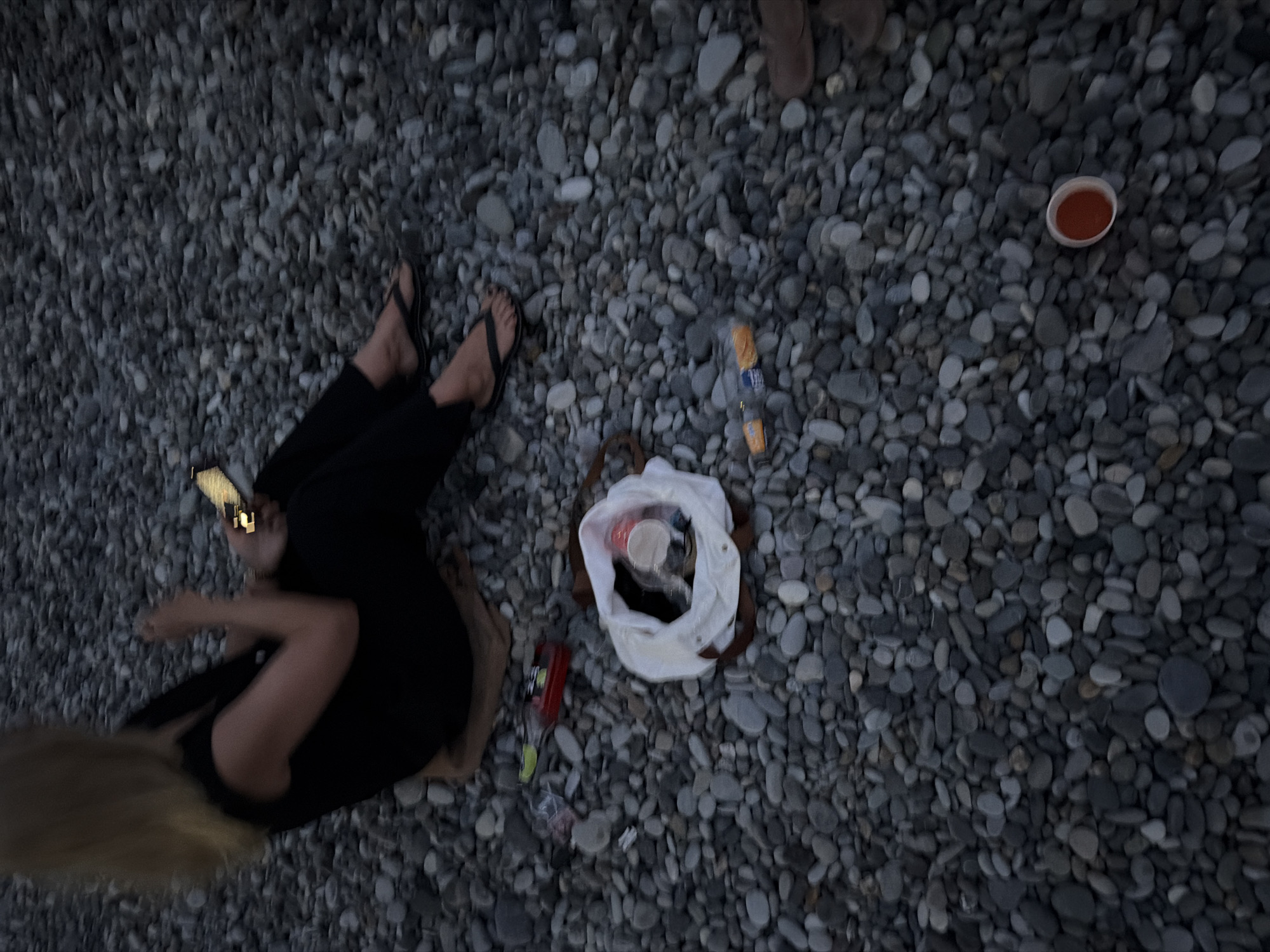 An individual sits on a pebble beach in Sochi, Russia, holding a mobile phone. The person is wearing a black sleeveless top and black pants, with black flip-flops on their feet. To their left, on the pebbles, are several items, including a red bottle with a green label, a white shopping bag containing a red cup and other unseen items, and two small orange cylindrical objects that appear to be wrappers. To the right of the bag, a single red cup with some reddish liquid is visible on the pebbles. The ground is entirely covered in gray and dark gray pebbles of various sizes.