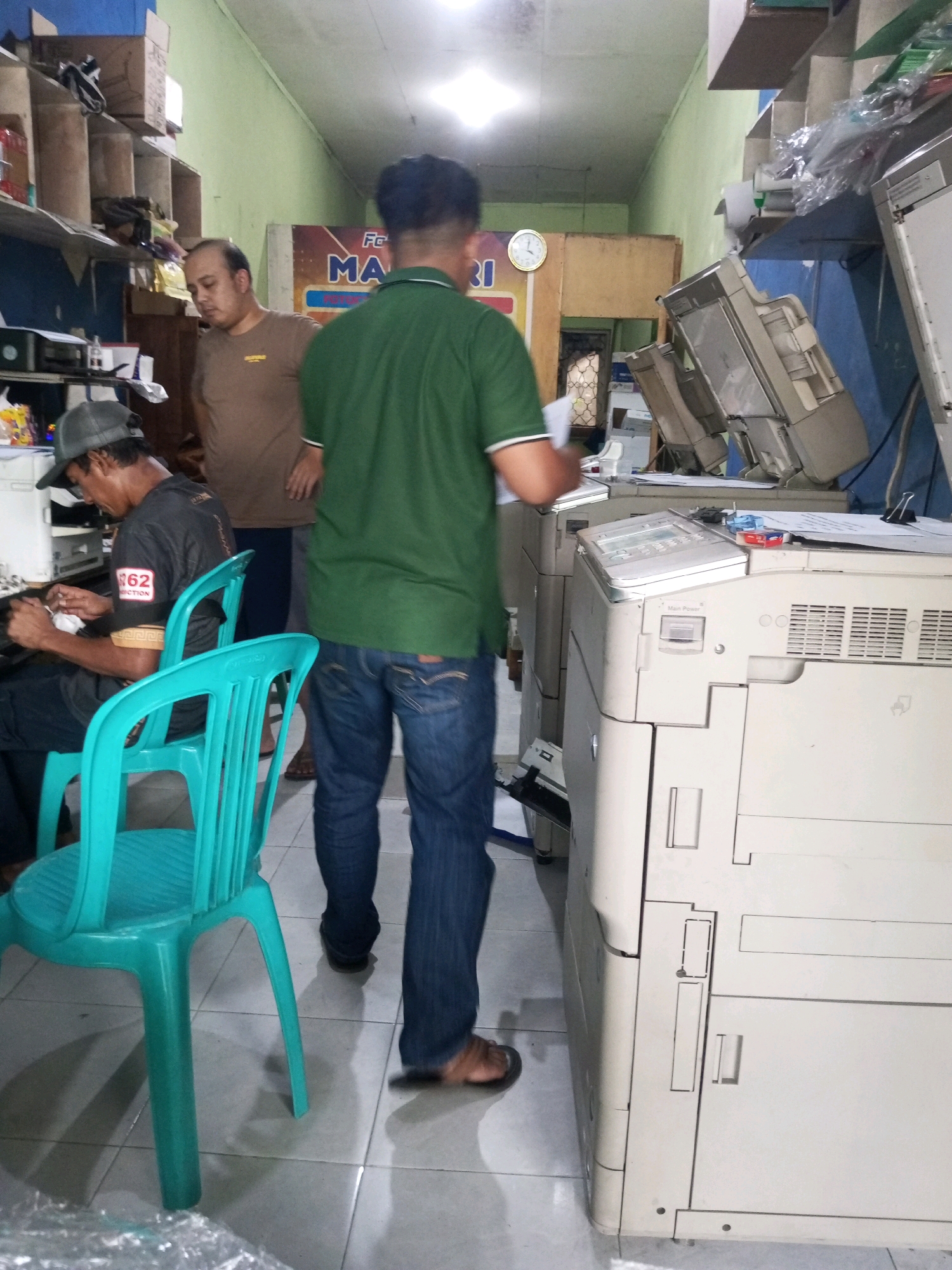The image depicts an interior scene, likely a small office or copy shop in Garung, Indonesia, filled with office equipment, predominantly large photocopiers. Three individuals are visible. In the foreground, to the left, a person wearing a grey baseball cap and a dark t-shirt with white lettering is seated and appears to be working with some papers or devices. To the right, a person in a green t-shirt and blue jeans is standing and facing away from the camera, looking towards the photocopiers. In the background, a man in a light brown t-shirt is standing and looking downwards. The environment is cluttered with stacked boxes on shelves along the left wall. Multiple photocopiers, some stacked, occupy the right side of the frame. The flooring is tiled. A clock is visible on the wall in the background, above a colorful sign with text.