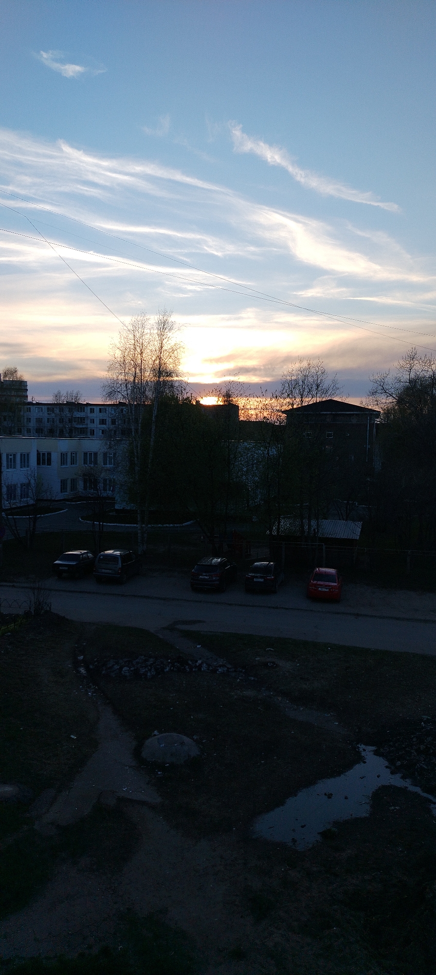 The image depicts an urban twilight scene. In the foreground, there is a dark, grassy area with visible ground and a possible puddle. A street separates this foreground from a row of parked cars of various models and colors, including black, red, and grey. Behind the cars are trees and buildings. A multi-story white and grey building is visible on the left, and a darker building with a sloped roof is on the right. The sky is light blue at the top, transitioning to orange and yellow at the horizon, indicating either sunrise or sunset. Wispy clouds are scattered across the sky. Power lines are visible in the upper part of the frame. The location appears to be a residential area within a city.