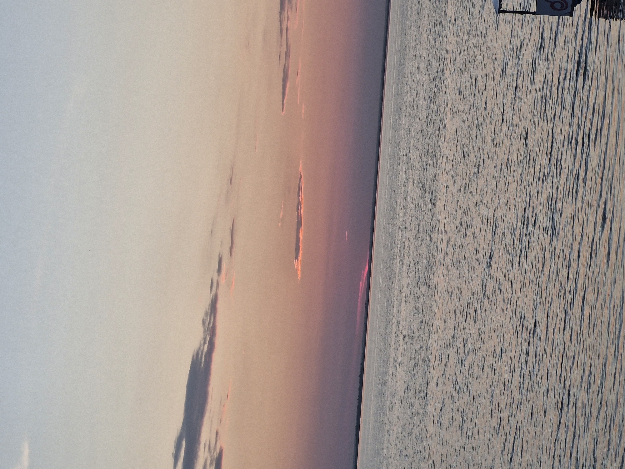 This image depicts a serene sunset over a body of water, likely part of Lake Kokchetav in Kokshetau, Kazakhstan. The sky transitions from a pale blue at the top to soft pinks and oranges near the horizon, with a few wispy clouds scattered across. The water is calm, reflecting the colors of the sky with gentle ripples. In the far distance, a faint landmass with some vegetation is visible along the horizon. On the right side of the image, the corner of a small, white boat or structure with a red logo is visible, partially submerged in the water. The overall atmosphere is tranquil and peaceful.