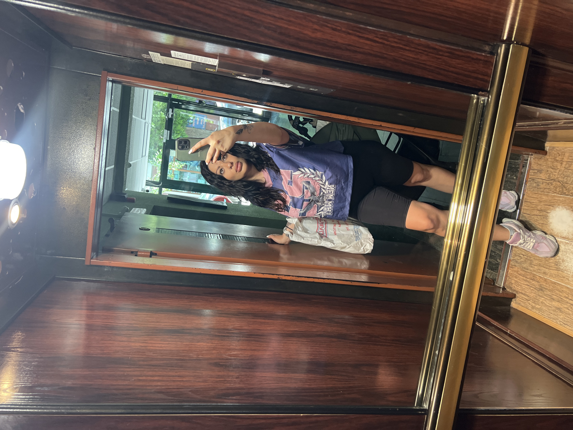 A woman stands in a wood-paneled elevator, taking a selfie with her phone. She is wearing a blue jersey, black shorts, and lilac sneakers. A white shopping bag rests on the elevator floor. The elevator is likely located in Plovdiv, Bulgaria. The outside of the elevator, visible in the mirror, suggests the elevator is opening into a city street.