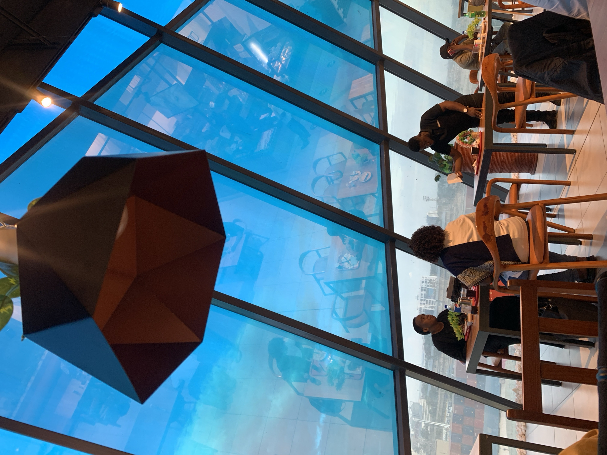 This is a photograph taken from inside a restaurant with a sloped, blue-tinted glass ceiling that offers a view of the city. The ceiling's angle creates a dynamic, angular effect. In the foreground, a large, dark, geometric pendant light hangs from the ceiling.
Below the ceiling, several tables are set up for dining. In the mid-ground, a man in a black polo shirt with a logo on the chest is serving a customer at a table. He is holding a small white box and appears to be placing it on the table. This table has a vase with flowers and a plate with food. Another table to the left has two women seated, one in a black top and another in a white top with a patterned overlay. They are looking in different directions, and there are drinks and plates on their table. Further to the right, another man is seated at a table, talking on a mobile phone, with a drink and some greenery on his table.
The view through the ceiling reveals reflections of the interior of the restaurant, including chairs and tables, superimposed on the exterior cityscape. Outside the restaurant, the cityscape shows buildings, and a large area with shipping containers, suggesting a proximity to a port. This view is characteristic of the Dar es Salaam port area, with its distinctive container terminal. The overall ambiance appears to be a modern dining establishment with a focus on a scenic view.