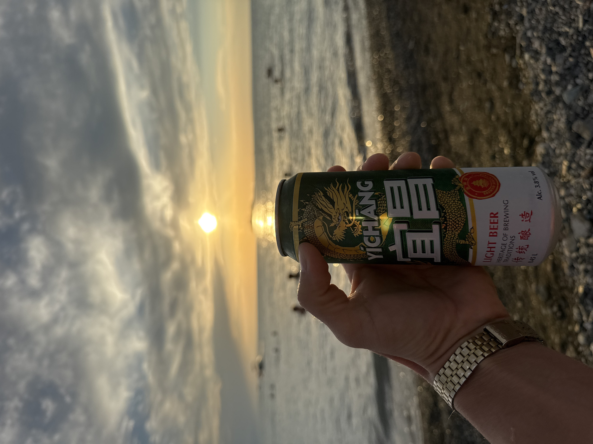 A close-up, eye-level shot shows a person's left hand holding a green and gold can of Yichang Light Beer. The can features a dragon illustration and Chinese characters. The background depicts a serene sunset over a body of water, likely the Urdzhar River in Urdzhar, Kazakhstan. The sun, partially obscured by clouds, casts a warm glow on the water's surface, creating shimmering reflections. Several indistinct figures can be seen in the water, appearing to be swimming or wading. The shore in the foreground is a mix of sand and pebbles. The person is wearing a gold metal watch on their wrist. The overall atmosphere is peaceful and relaxed.