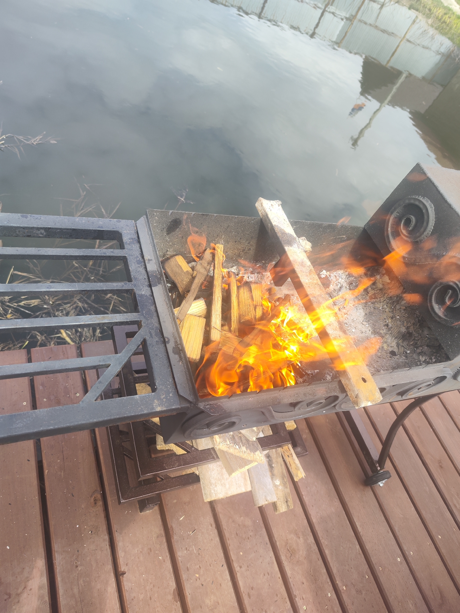 Here is a detailed description of the image:
**Scene:** The image shows a metal grill with wood burning inside, positioned on a wooden deck. Adjacent to the grill is a body of water, possibly a lake or river, reflecting the sky and some distant structures.
**Objects:**
* **Grill:** The grill is made of metal, with decorative spiral designs on the side. It contains burning wood and ash.
* **Wood:** There are pieces of wood both burning inside the grill and stacked underneath it on the deck.
* **Deck:** The deck is made of wooden planks.
* **Water:** A body of water is adjacent to the deck, showing reflections.
* **Fence:** A partial view of a fence is visible in the background, across the water.
**Actions:**
* Wood is actively burning inside the grill, producing flames.
**Environment:** The location appears to be outdoors, next to a body of water, possibly in a backyard or recreational area.
**Location:** The city is Unknown, Unknown.