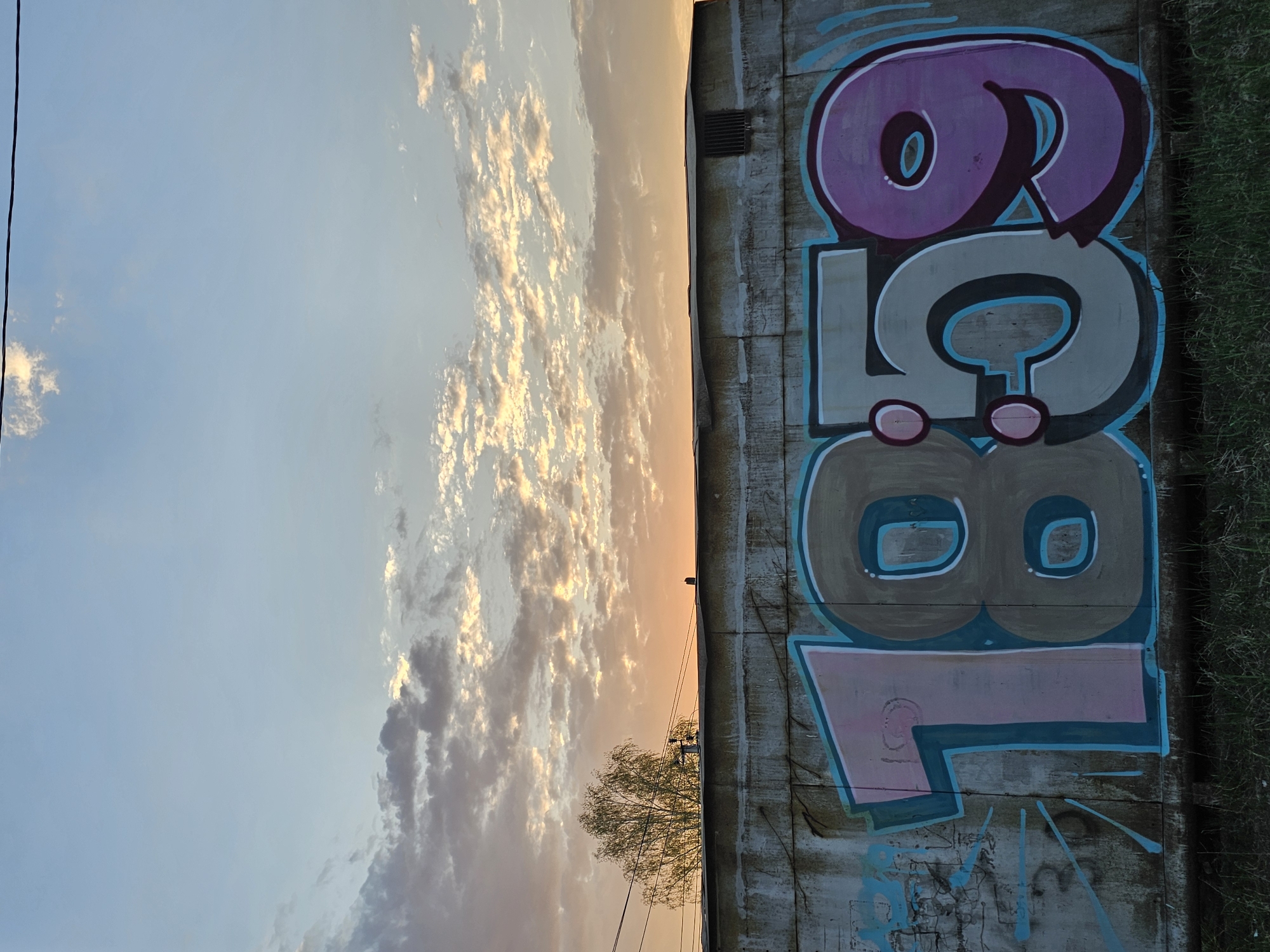 A graffiti-covered concrete wall stands against a backdrop of a sunset sky. The graffiti consists of large, stylized numbers in shades of purple, grey, and brown, outlined in teal. The sky is a gradient of blues and oranges with scattered clouds. To the left of the wall, there's a power line and a tree silhouetted against the sunset. The image is taken during sunset. The location is an unknown city and state.