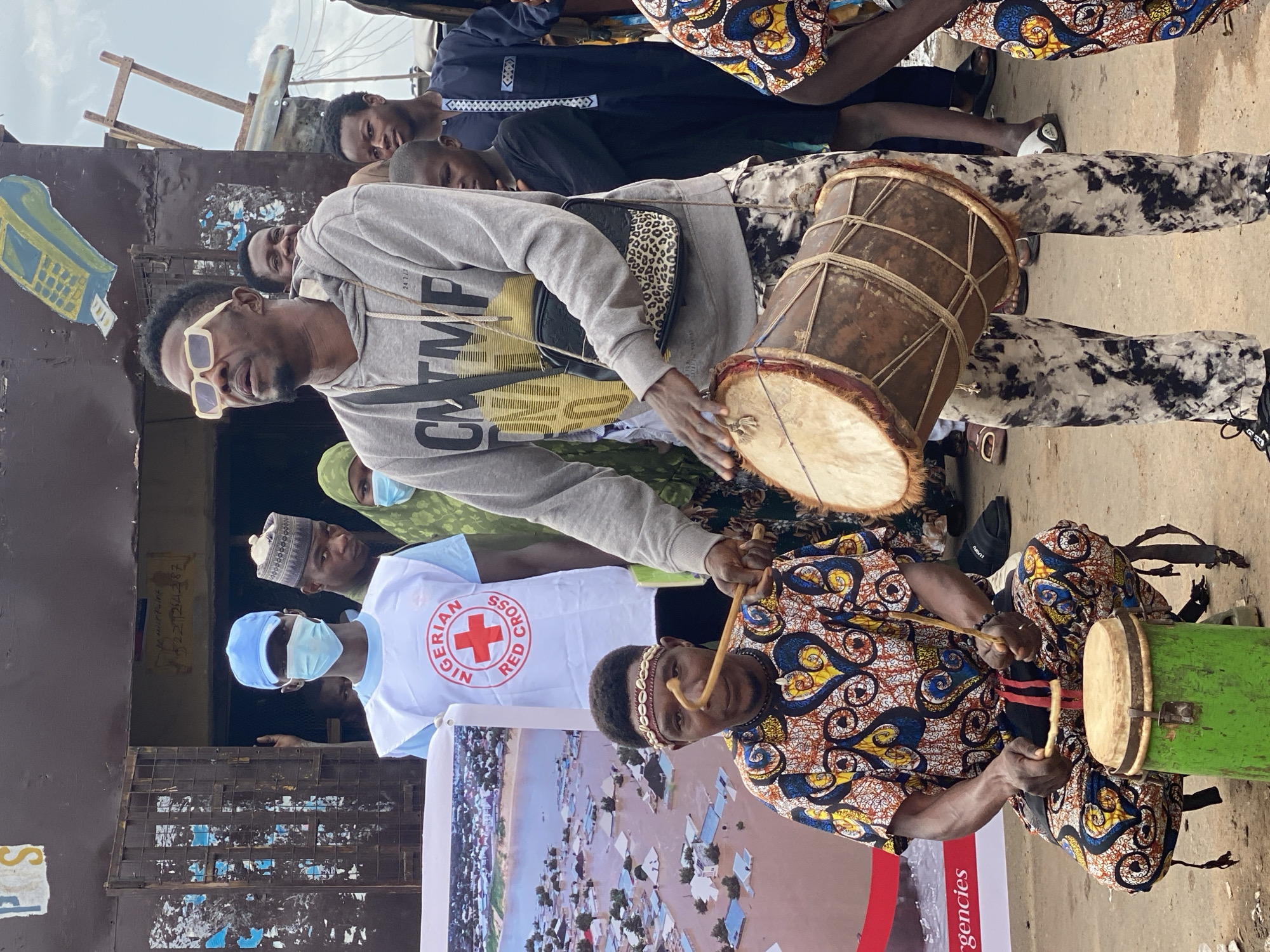 A group of people are gathered outdoors in front of a weathered building in Rigachikun, Nigeria. In the foreground, a man with a beaded headband is playing a small green drum with sticks, dressed in a vibrant, patterned traditional outfit. To his right, another man, wearing sunglasses and a grey hoodie with a leopard-print crossbody bag, is playing a larger, brown drum. Behind them, a person in a white vest with a Red Cross logo and a blue face mask is holding a large poster depicting aerial views of flooded homes. Other individuals, some wearing face masks and traditional attire, stand and observe the scene. The ground is dirt, and debris is visible. The building behind them appears to be a shop or storefront with a dark, corrugated metal facade and visible graffiti. The sky is overcast.