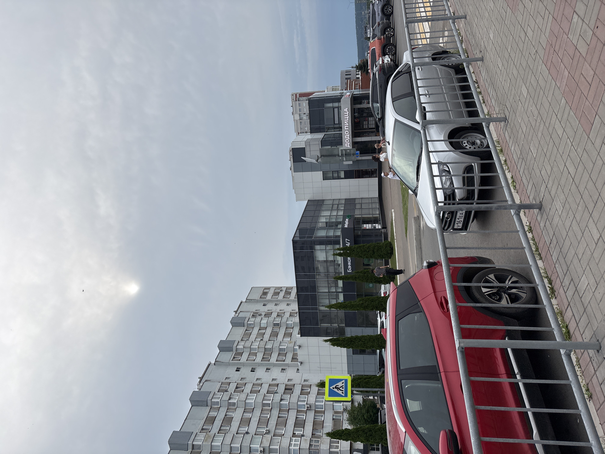 A street scene in Naberezhnye Chelny, Russia, features a multi-story residential building on the left and a modern commercial building with a "Dodo Pizza" sign on the right. A silver sedan with license plate Y161EA is parked in front of the commercial building, while a red SUV is parked closer to the foreground, separated by a metal railing. Several people are visible walking near the commercial building, and cars are parked in the background. The sky is overcast with the sun faintly visible.