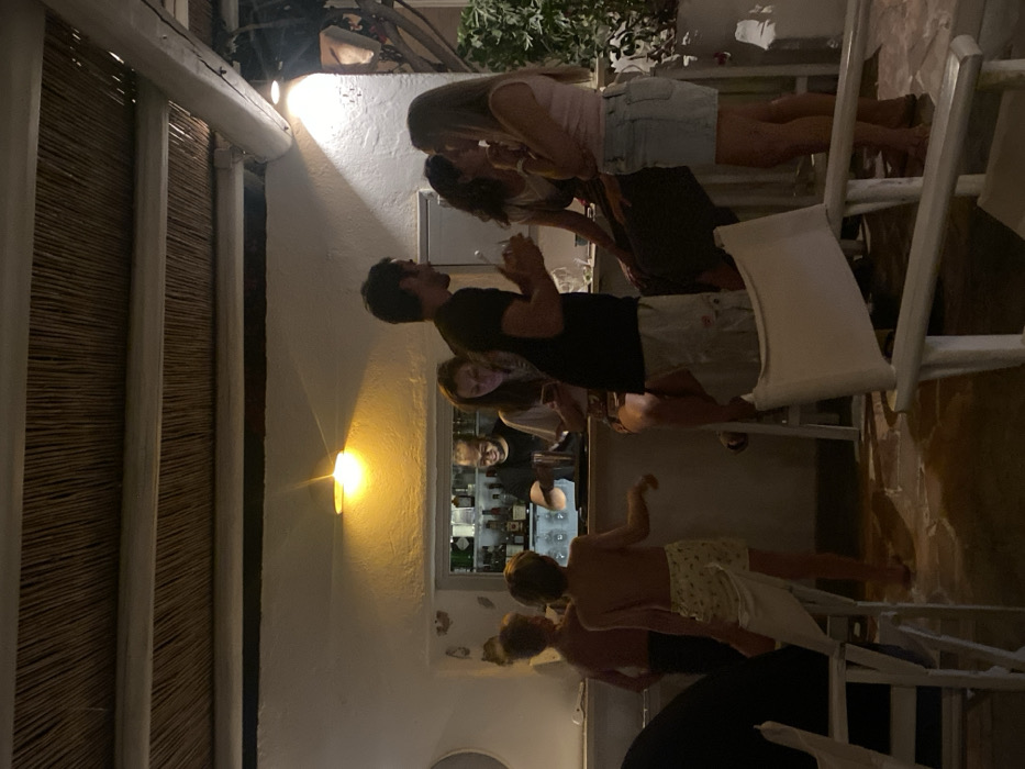 The image depicts an evening scene within a bar or restaurant in Φάρος, Greece, characterized by white-washed walls and a thatched ceiling with white wooden beams. Nine individuals are visible. At the center, a male bartender with a beard, wearing a dark shirt, is positioned behind a well-lit bar counter, actively mixing a drink. Behind him, shelves are stocked with bottles and glassware. To the right of the bartender, a female is seated at the bar, observing a mobile phone. Adjacent to her, a standing male holds a glass and a stirrer, facing two seated females. The female on the left has dark hair, while the female on the right, wearing a light pink top and a light denim skirt, has blonde hair; both are engaged in conversation with the standing male. In the left foreground, two shirtless young boys are standing near the bar area; one, closer to the viewer, wears patterned shorts and faces away, partially obscured by a white chair. The flooring is light-colored and appears to be stone or tile. White-painted wooden furniture, including bar stools and chairs, is present. Green foliage is visible in a planter on the right side of the frame.