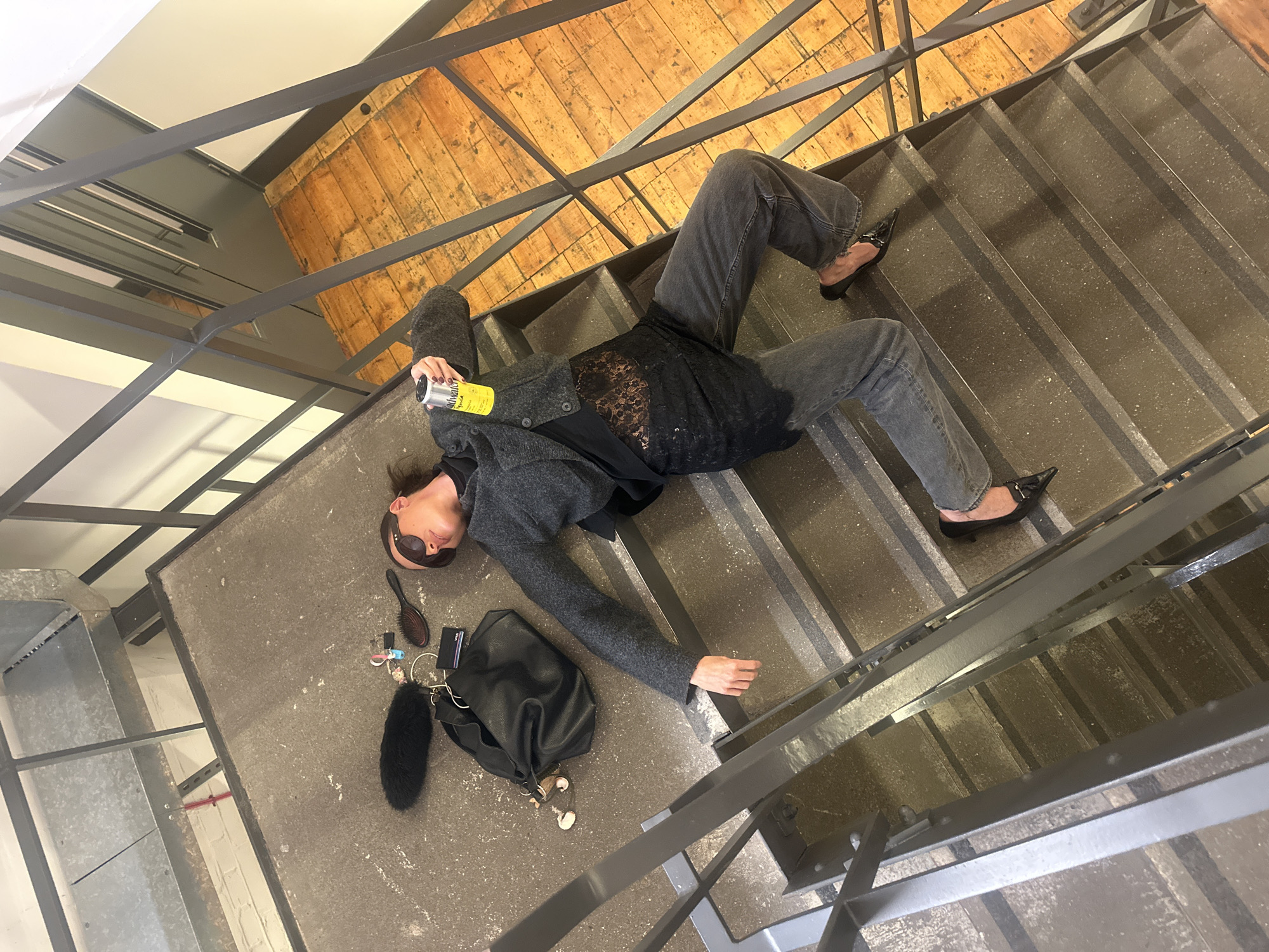 An overhead shot captures a person lying sprawled across a concrete staircase and landing within what appears to be a modern or industrial-style building. The individual, dressed in a dark grey cardigan or coat over a black lace-detailed top and dark grey jeans, is positioned diagonally, with their head near the landing and their legs extending down the steps. They wear black pointed-toe flat shoes and dark sunglasses.
In their right hand, the person holds a yellow can, partially obscured, but "saltwater" and "Spindri" (likely Spindrift) are discernible on the label. Scattered on the concrete landing near their head are personal items: a black hairbrush, a black leather handbag, a set of keys with multiple keychains including a small plush toy, and a black furry object, possibly a microphone wind muff or a bag charm.
The staircase features dark grey concrete steps and risers, with black metal railings forming the banister. In the upper part of the image, a wooden plank floor is visible at the next level, suggesting an upper story or platform. Overhead, metal beams and a white ceiling are seen. The lighting suggests an indoor daytime setting. The overall scene, located in London, United Kingdom, presents an ambiguous narrative, possibly staged, implying anything from rest to a stumble.