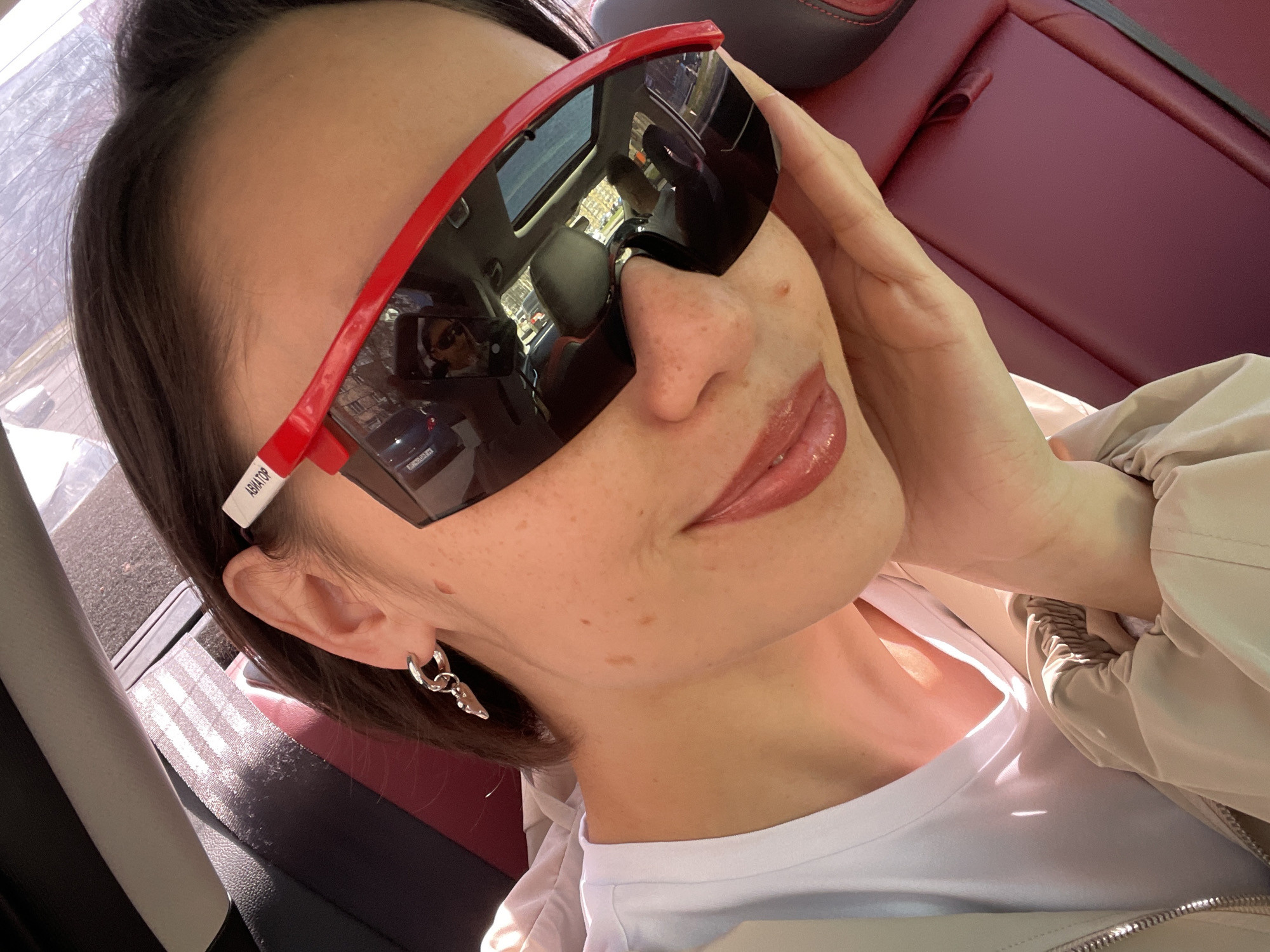 A woman is posing for a selfie while in a car. She is wearing large, red-framed sunglasses that obscure much of her face, a white t-shirt, and a light-colored jacket. Her hair is dark and short. She has freckles and is wearing lip gloss. The car interior features red leather seats. The sunglasses reflect the interior of the car and a view through the sunroof, including buildings and possibly another person. The location is likely Izhevsk, Russia, based on the text "AVIATOR" on the side of the sunglasses.