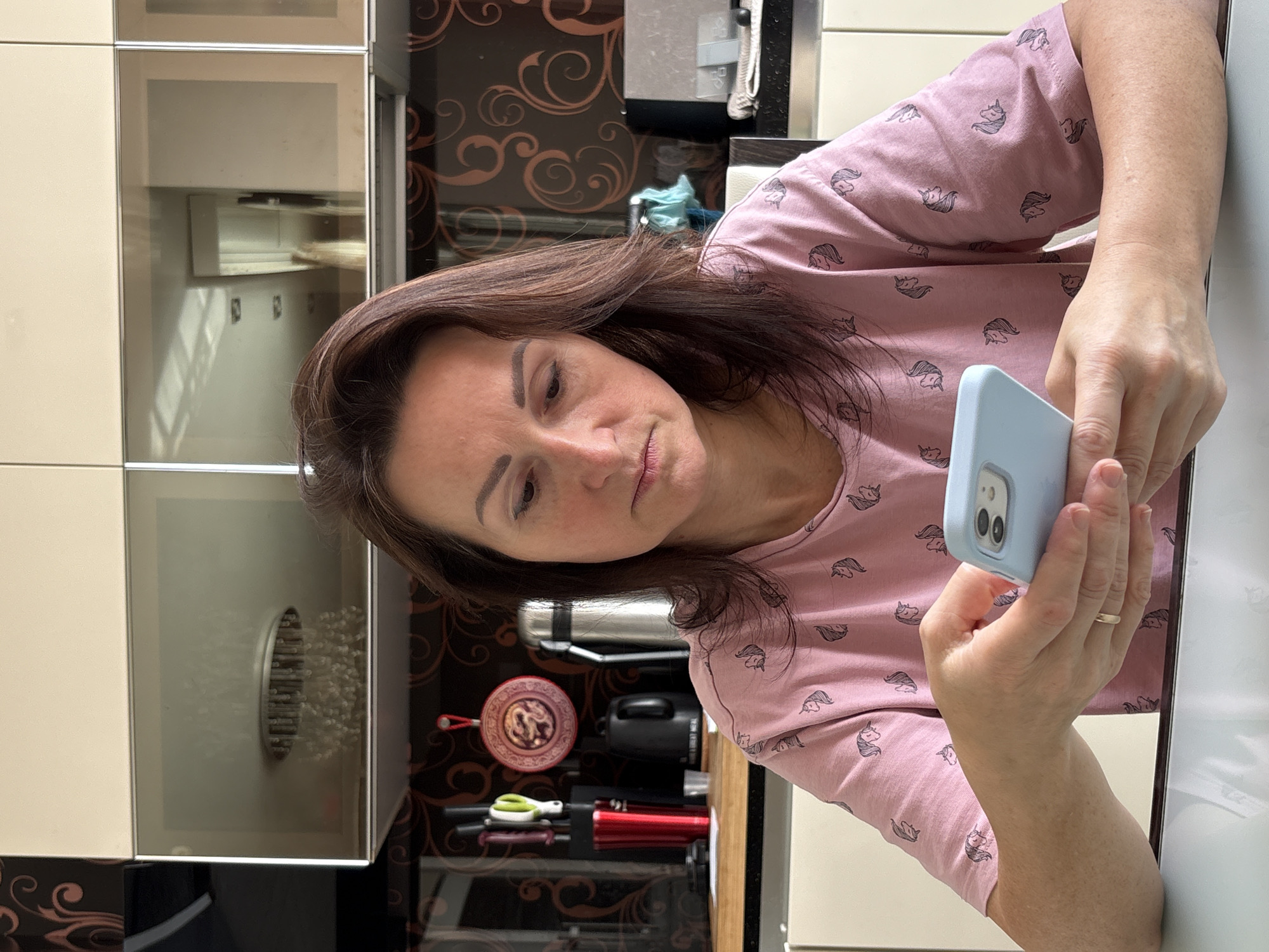 An indoor, eye-level shot captures a woman engrossed in her smartphone in a kitchen setting in Saint Petersburg, Russia. She has dark brown hair and is wearing a pink long-sleeved top patterned with small unicorn heads. She holds a light blue-cased smartphone in both hands, gazing at its screen with a focused, somewhat contemplative expression. A gold ring is visible on her left ring finger.
The background reveals a modern kitchen. To her left are light, possibly beige, upper cabinets with reflective surfaces. Behind her, a dark brown or black wall or backsplash panel features ornate, glossy brown or gold swirling patterns. On the counter below this panel, several objects are arranged: a silver thermos or kettle, a small red decorative pan with an intricate design, a black appliance, a red bottle or container, and a pair of green-handled scissors. The white surface where her arms rest appears to be a countertop, also exhibiting reflections of the room. The overall lighting suggests it is daytime. There is no visible text in the image.