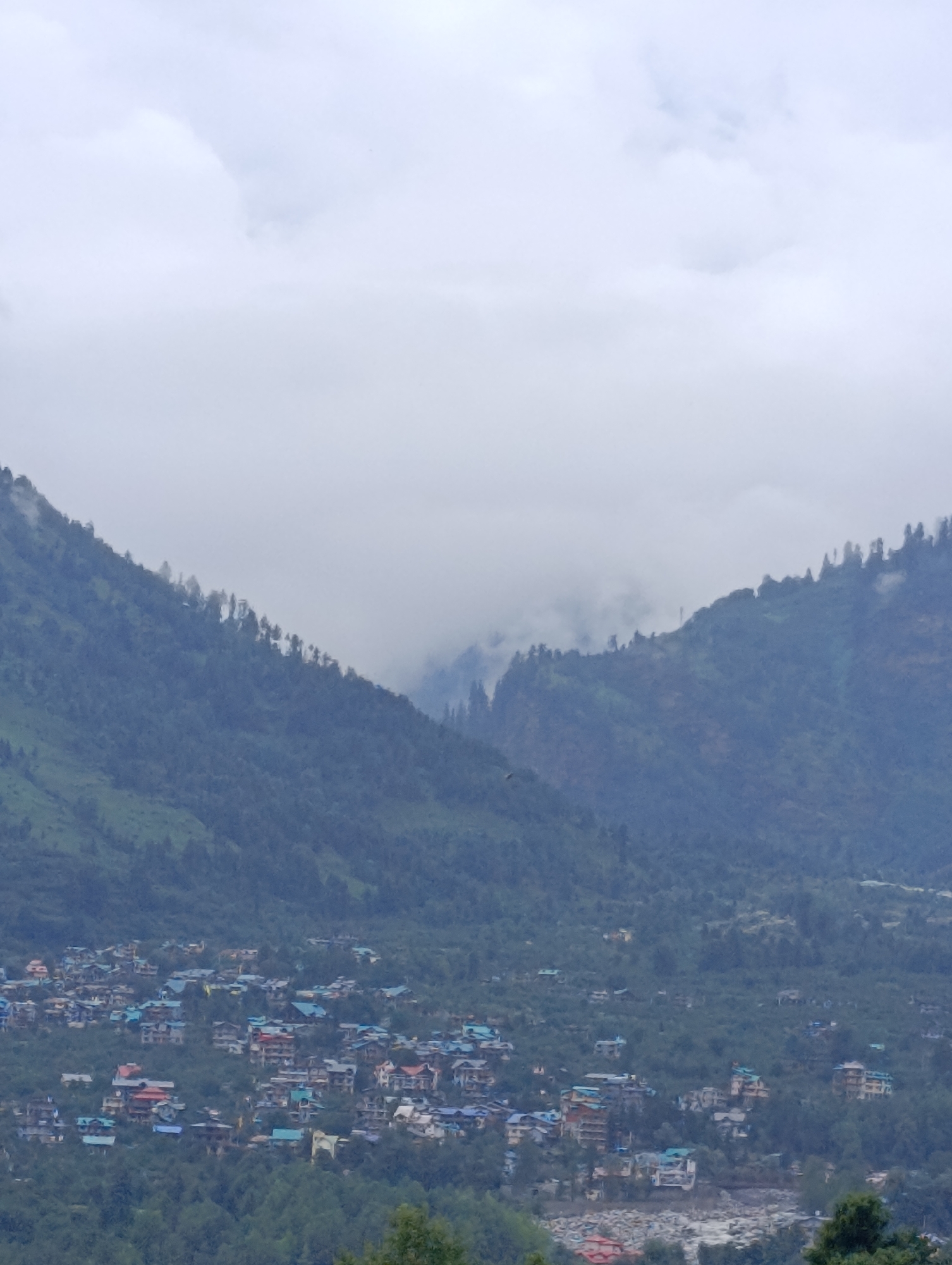 The image displays a panoramic view of Vashisht, India, a town nestled in the Himalayas. The scene is dominated by lush green mountains, partially obscured by a thick blanket of clouds. A settlement, characterized by a cluster of multi-story buildings with colorful roofs, is visible on the lower slopes of the mountains. The buildings appear to be residential or commercial structures, and their varied hues create a vibrant contrast against the verdant landscape. A riverbed, filled with stones, can be seen in the lower right portion of the image, suggesting the presence of a water body. The overall atmosphere is serene and picturesque, indicative of a mountain town in a cloudy, possibly monsoon, season.