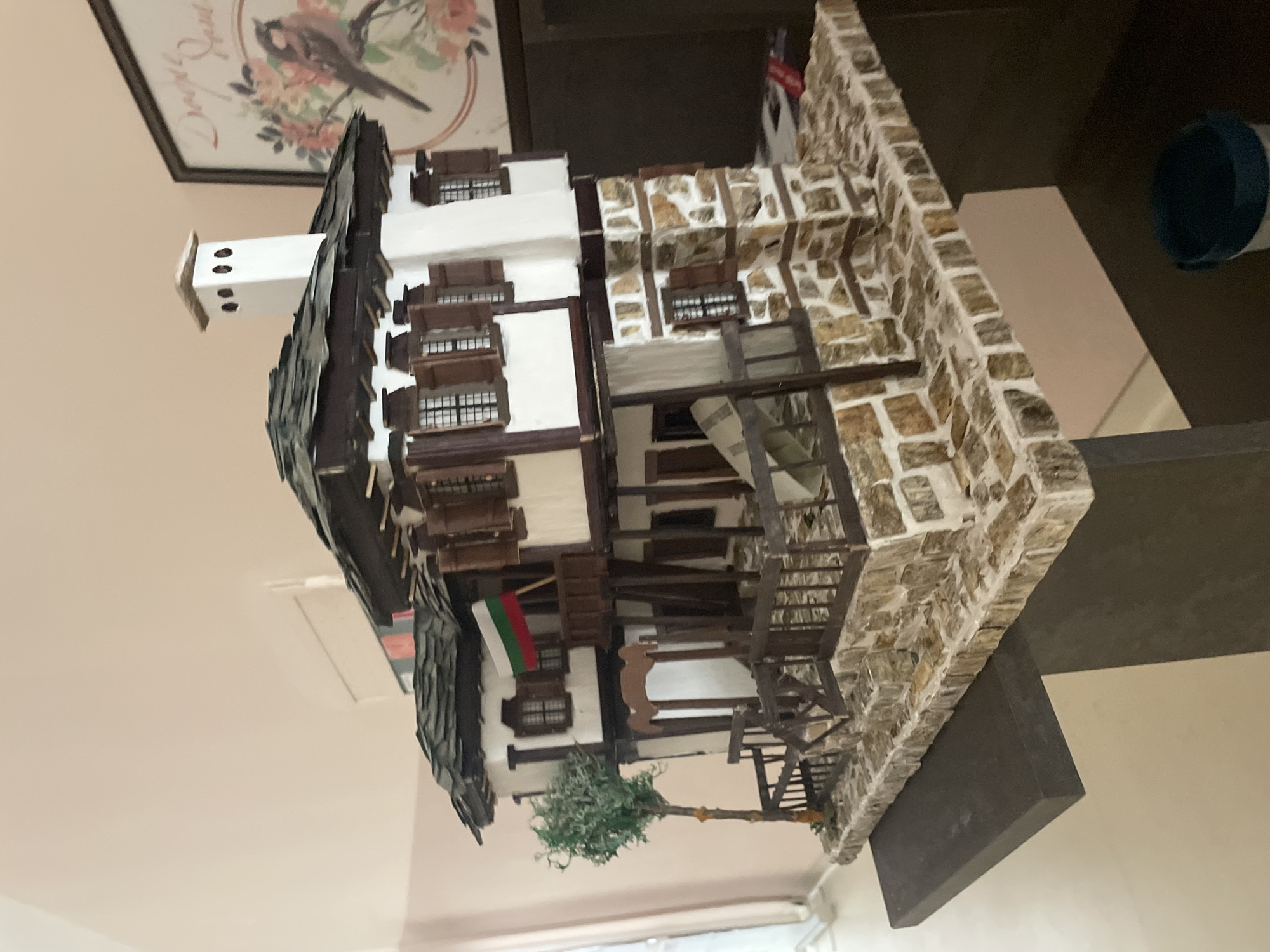 The image depicts a miniature model of a traditional Bulgarian house, likely a replica of one found in Zlatograd, Bulgaria. The house features white walls, dark brown wooden trim and window frames, a stone façade on one side, and a steeply pitched roof made of dark grey tiles. A small Bulgarian flag is displayed on one of the upper floors. A miniature tree is placed at the base of the model. The model sits on a dark grey shelf. In the background, there is a framed picture and a glimpse of the room it's in. The scene appears to be indoors, possibly in a private residence.
