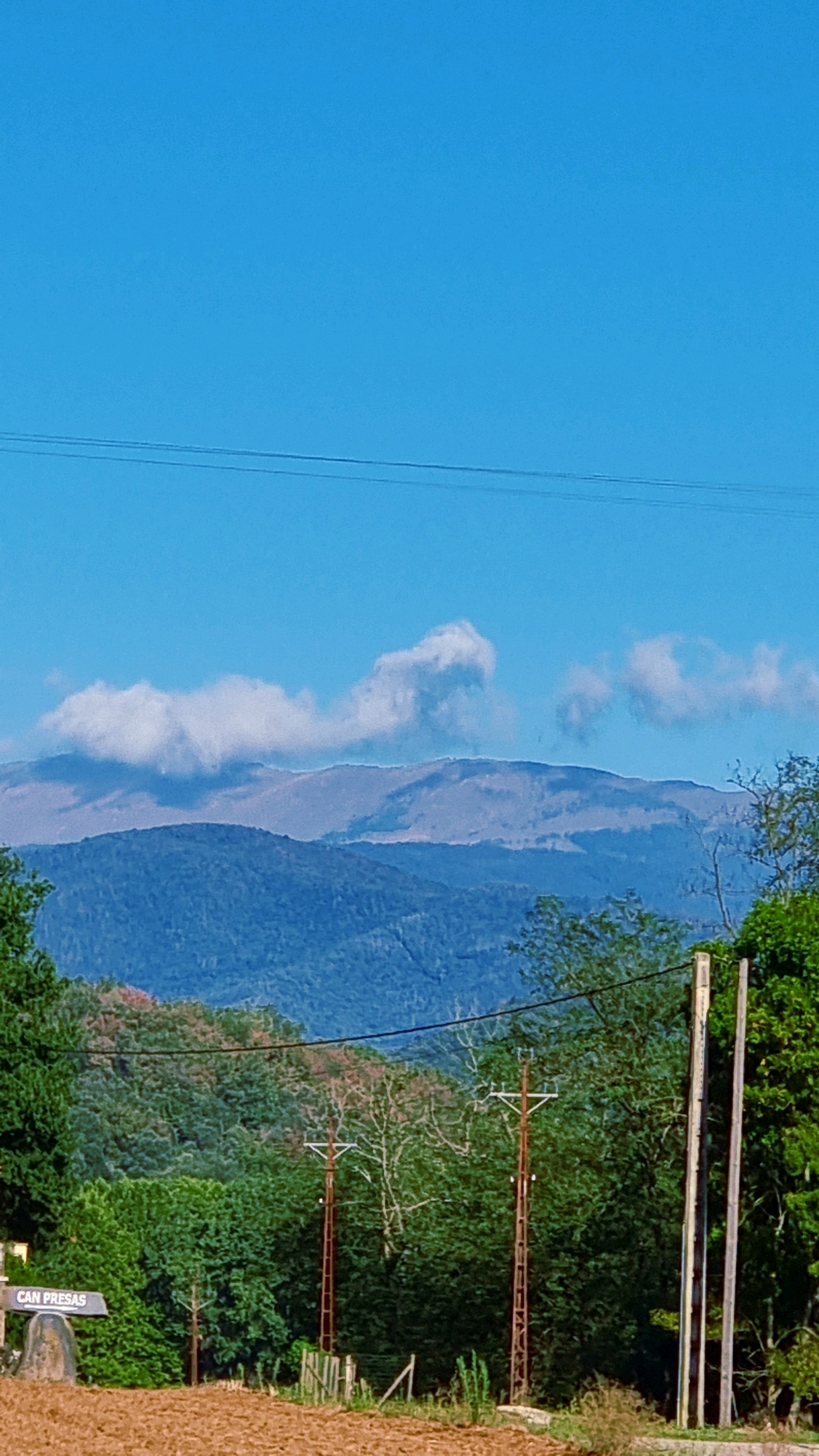This is a vertical, eye-level outdoor photograph of a mountainous landscape under a clear blue sky. In the foreground, a patch of brown soil is visible, with some dry grass and a small section of a wooden fence. To the left, a sign with the text "CAN PRESA" is mounted on a rough stone base. Behind the soil and fence, a dense line of green trees stretches across the middle ground, with some lighter foliage and bare branches intermixed. Two tall, rusty metal utility poles with crossbars are prominent in the middle ground, with a third, lighter pole visible to the right.
The background consists of layered mountains. The closest mountains are covered in dark blue-green foliage, becoming progressively lighter and more muted in color with distance. The furthest mountain range appears hazy and light brown, with scattered white clouds partially obscuring its crest. Two thin, dark horizontal lines, presumably power lines, stretch across the upper portion of the sky. The scene appears to be a rural or semi-rural area, likely in the municipality of Llocalou, Spain, given the sign. No people are visible in the image.