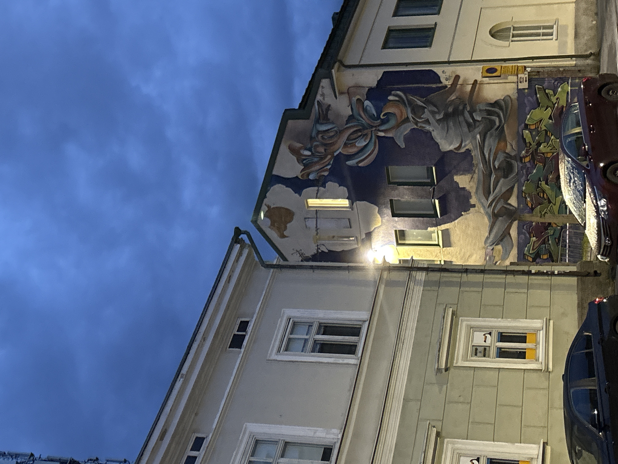 In the city of Pori, Finland, a nighttime scene captures the facade of two adjacent buildings. The building on the left is painted a light green with white trim around its windows. The building on the right is adorned with a large, vibrant mural depicting an abstract, flowing form in shades of blue, orange, and gray. The mural covers a significant portion of the building's exterior. A streetlamp casts a yellow glow on the scene. The sky above is a deep blue, filled with dark, ominous clouds. A telecommunications tower is visible on the far left, partially obscured by the building. Two cars are parked on the street in front of the buildings; one is dark, and the other is a reddish-maroon color with a dusting of snow on its hood. The street appears to be paved.