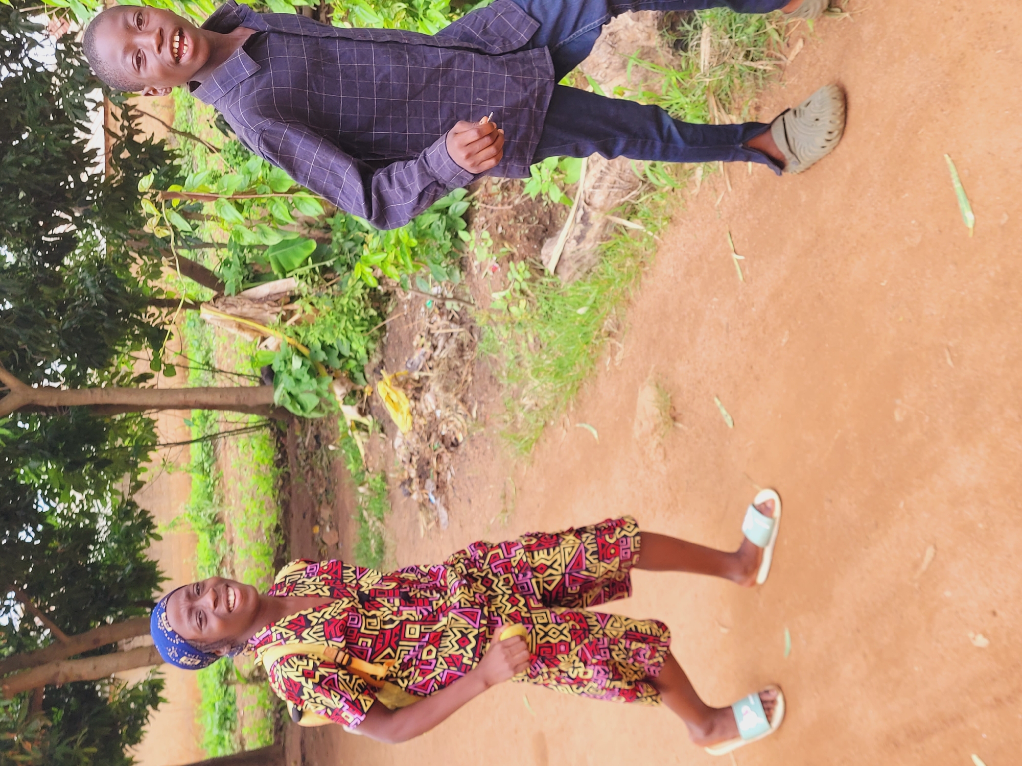 This image features two individuals, a young boy and an older woman, standing on a reddish-brown dirt path in an outdoor setting, likely in Bukuru, Nigeria. The scene is bright, indicating daytime and clear weather.
The young boy, positioned towards the top left of the frame (when viewed upright), has dark skin and short dark hair. He is smiling widely, looking directly at the camera. He wears a dark blue or black long-sleeved shirt with a subtle checkered pattern, dark blue trousers, and grey slip-on shoes or clogs. In his right hand, he holds a small, light-colored object.
The woman, located towards the bottom right of the frame, also has dark skin and is smiling towards the camera. She wears a blue and white patterned headscarf and a brightly colored short-sleeved top and matching shorts (or a dress) adorned with vibrant red, yellow, black, and white geometric patterns. A yellow strap, possibly from a bag, is visible over her shoulder. She is holding a yellow fruit, resembling a small banana or plantain, in her right hand and wears light blue and white sandals.
Both subjects appear relaxed and joyful, suggesting they are posing for a photograph. The background is rich with green vegetation, including numerous trees with broad leaves and dense undergrowth, indicating a lush environment consistent with a rural or semi-urban area. There is no visible text in the image.
