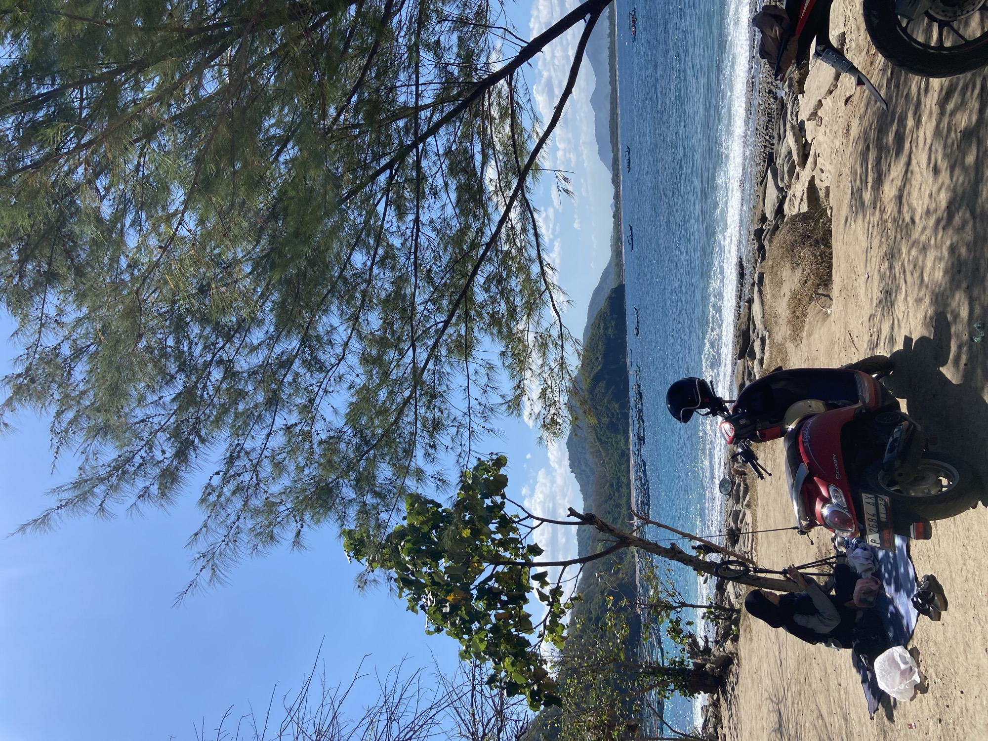 A coastal scene in Unknown, Indonesia, with a red scooter parked on a sandy area beside the water. A person is sitting next to the scooter, and a helmet rests on it. The ocean is visible with small boats in the distance, and the coastline is lined with trees. The sky is blue with some clouds, partially obscured by the branches of trees in the foreground.