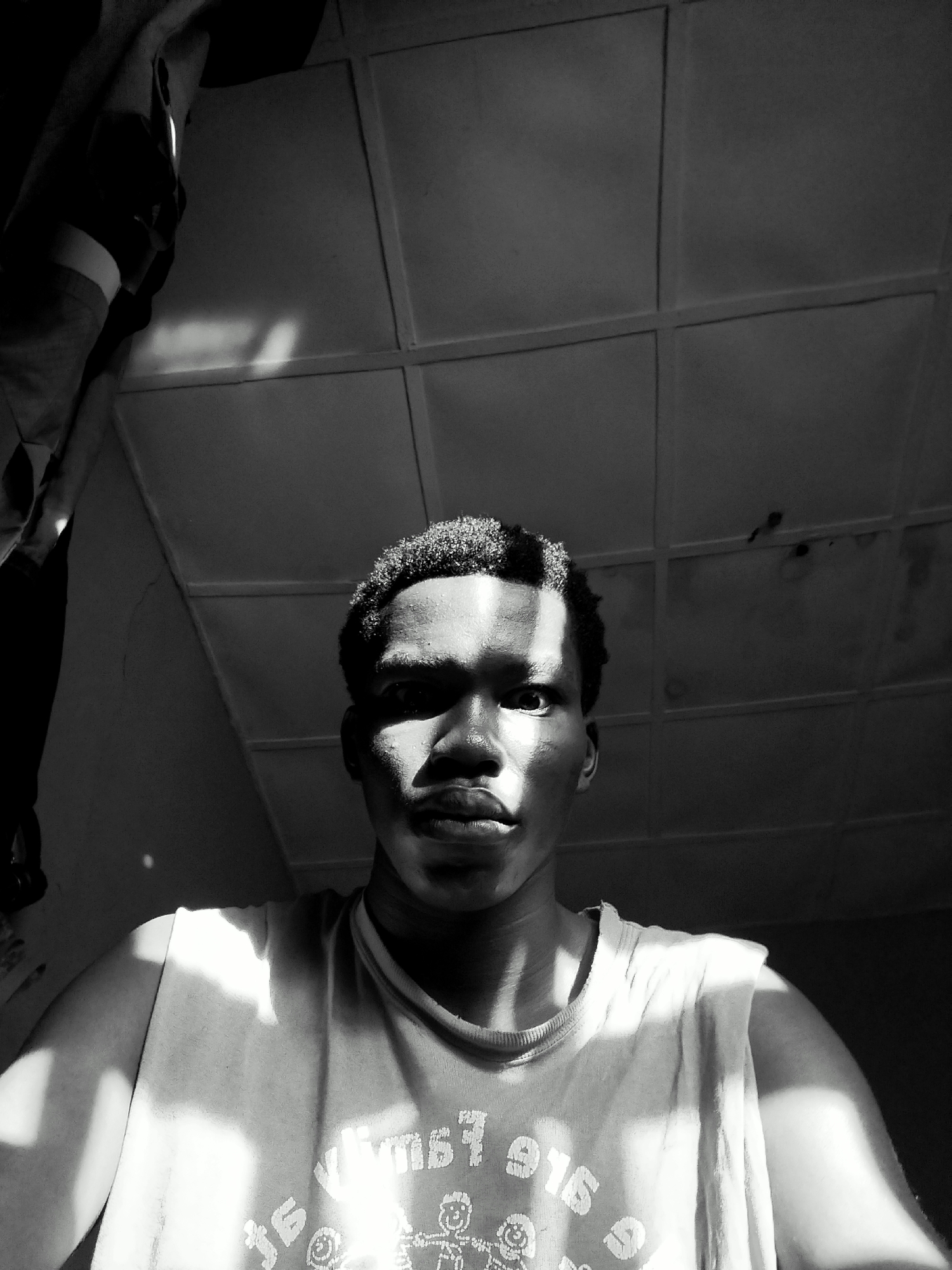 A black and white, high-contrast, close-up portrait shows a young Black man with short hair wearing a tank top. Light streams from an unseen source, dramatically illuminating one side of his face and his torso while casting deep shadows on the other side. The background consists of a ceiling with a grid-like pattern and some shadowed items hanging in the upper left corner. The location is indicated as Keffi, Nigeria, but there are no visual clues in the image to confirm this.