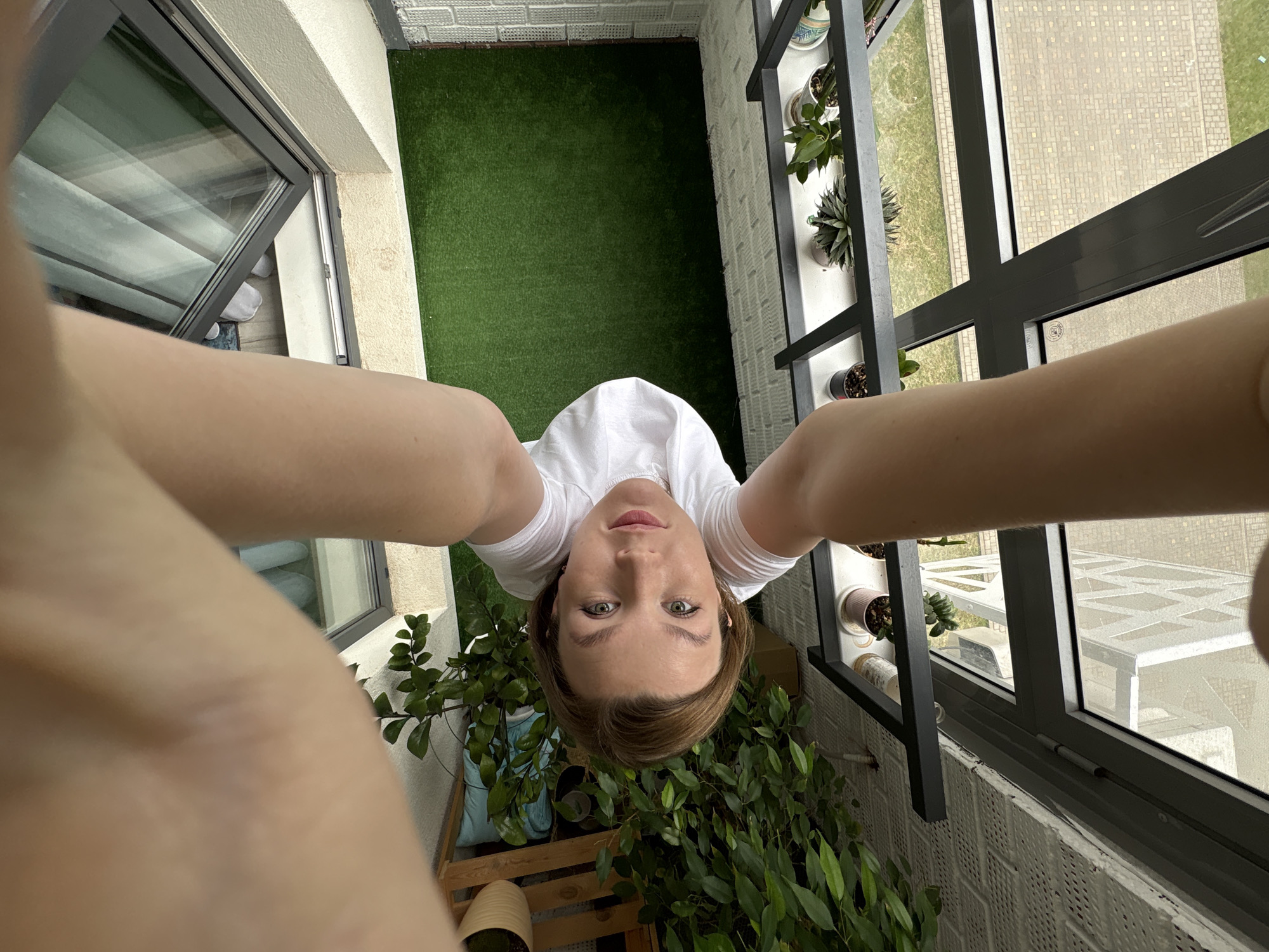 The image is a selfie taken from a high angle, looking down at a young woman with shoulder-length brown hair and green eyes. She is wearing a white t-shirt and is standing on a balcony with artificial turf. There are several potted plants on the balcony, including a large leafy plant to her left. A glass door and window are visible behind her, reflecting the sky and some interior elements. The lower part of the image shows the woman's arms and hands holding the camera. The scene appears to be a residential balcony, possibly in a city. There is no information available to confirm the location as Izhevsk, Russia.