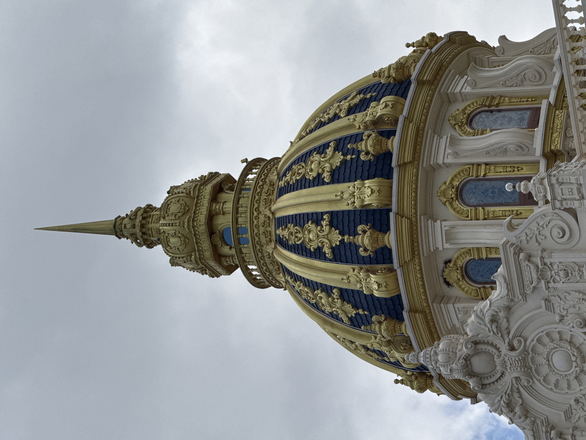 The image shows a close-up of a golden dome topped with a spire. The dome has dark blue tiles and ornate gold detailing. Below the dome, a white, intricately carved structure with arched windows is visible. The sky above is overcast with some patches of blue. This is the exterior of the Dinh 1 (also known as Bach Dinh or Villa Dupont) in Da Lat, Vietnam. There are no people or actions depicted in the image.