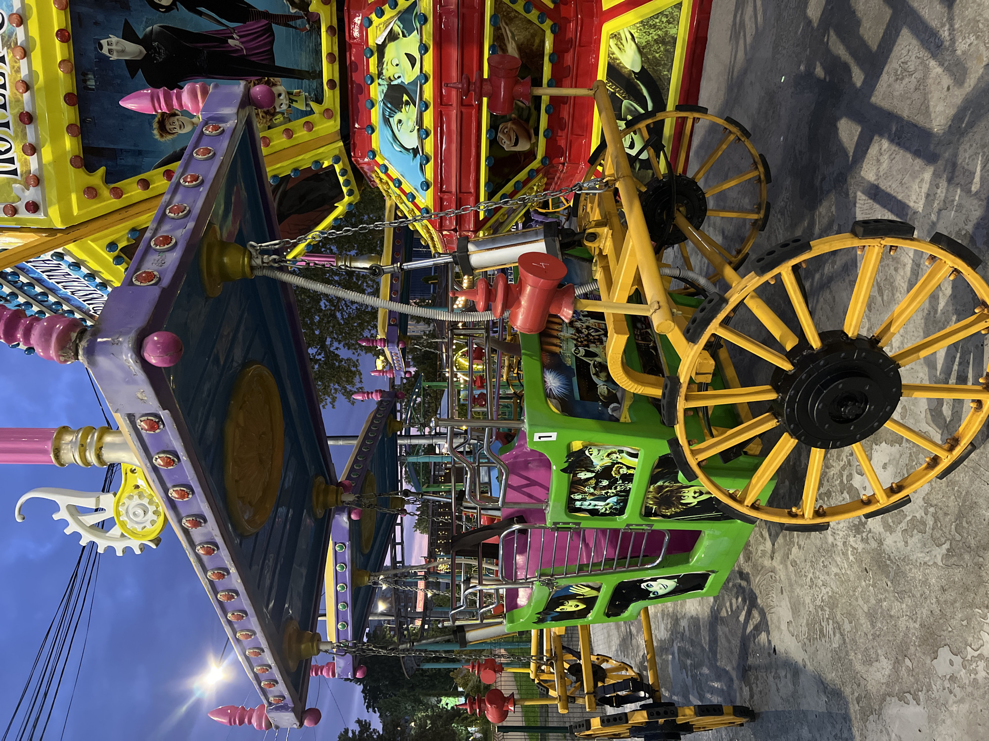 The image shows a close-up of a colorful amusement park ride. The ride is decorated with cartoon characters from the Hotel Transylvania franchise. The ride has a green carriage with yellow wheels and a purple canopy. In the background, there is another part of the ride that is red and yellow, also decorated with cartoon characters. The sky is dark blue, suggesting it is dusk or early evening. The ground appears to be concrete or asphalt. There are no people visible in the image. The location is identified as the "Hotel Transylvania" amusement ride in the city of Krasnodar, Russia.