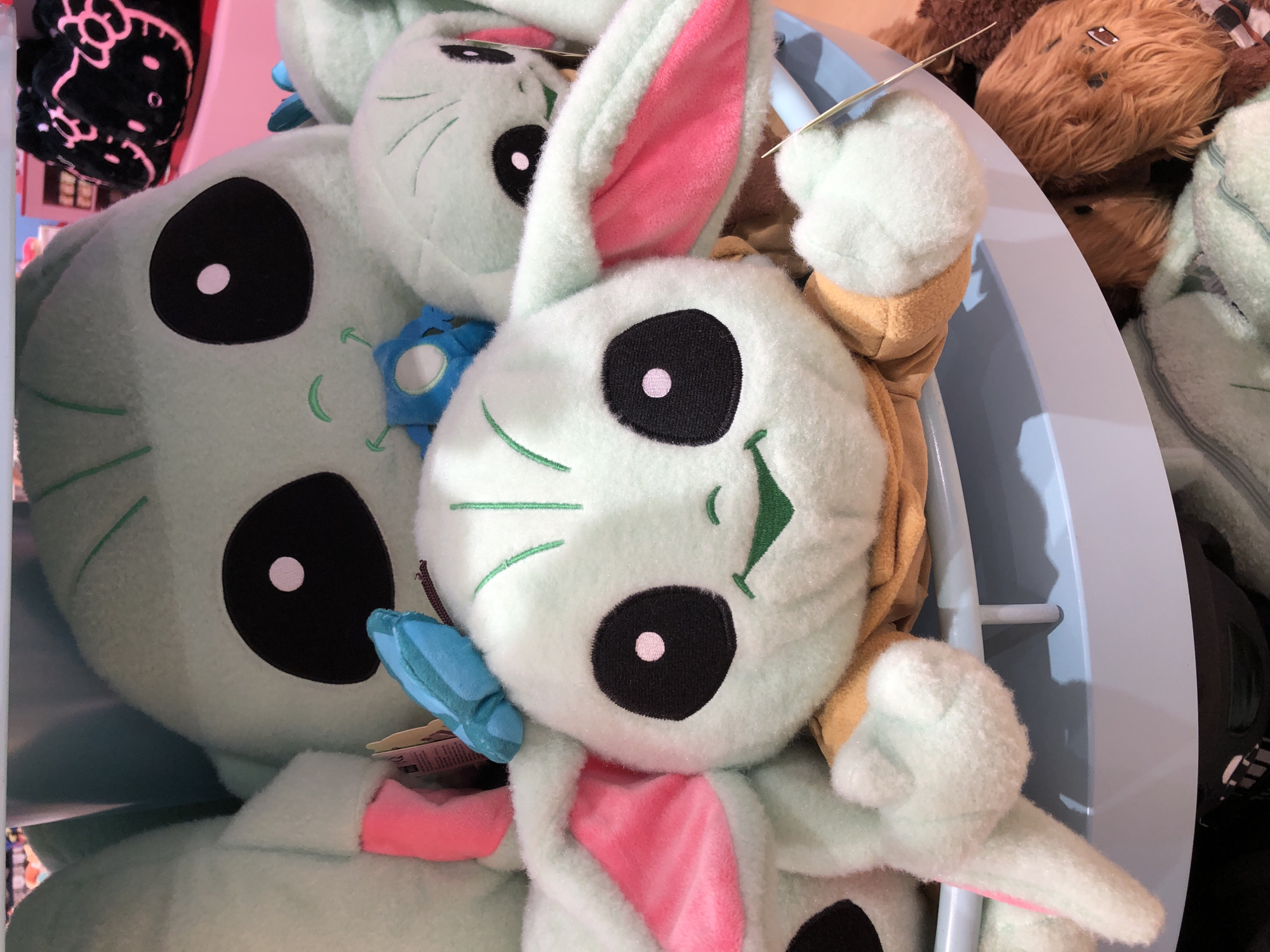 This media file features a close-up shot of several plush toys, primarily in pastel green and light blue, with accents of pink and tan. The main subject is a light green plush toy with large black eyes that have pink pupils, long floppy ears lined with pink, and green stitched facial features, including whiskers and a small nose. This toy appears to be in a seated position, with its arms outstretched.
Behind this toy, another similar light green plush toy is visible, also with large black eyes and green stitched features. This second toy has a blue butterfly-shaped accessory attached to its chest. To the right of the main subjects, a fuzzy brown plush toy, possibly a bear or a creature from Star Wars, is partially visible.
The toys are arranged in what appears to be a display basket or bin, indicated by the blue plastic edges and the reflective surface of the bin. In the background, on the left, a black plush blanket with a pink Hello Kitty design is visible, suggesting a retail environment.
There is no discernible time of day or weather information. The setting appears to be indoors, likely a store, given the display of merchandise. The emotions conveyed are playful and childlike, due to the nature of the plush toys.
Visible text includes a tag on the blue accessory of the rear plush toy, which is largely unreadable but contains some printed lines.