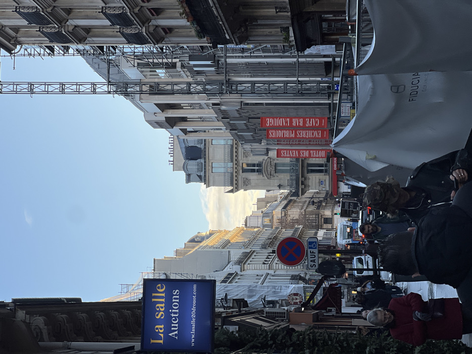 An urban street scene features buildings with scaffolding and signs. A blue sign reads "La salle Auctions." Red banners advertise "HOTEL DES VENTES," "ENCHERES PUBLIQUES," and "CAFE BAR L'ADJUGE." A "FIDUCIA" umbrella is present. A no parking sign with a disabled symbol is visible. People are walking on the street, adding to the bustling atmosphere.