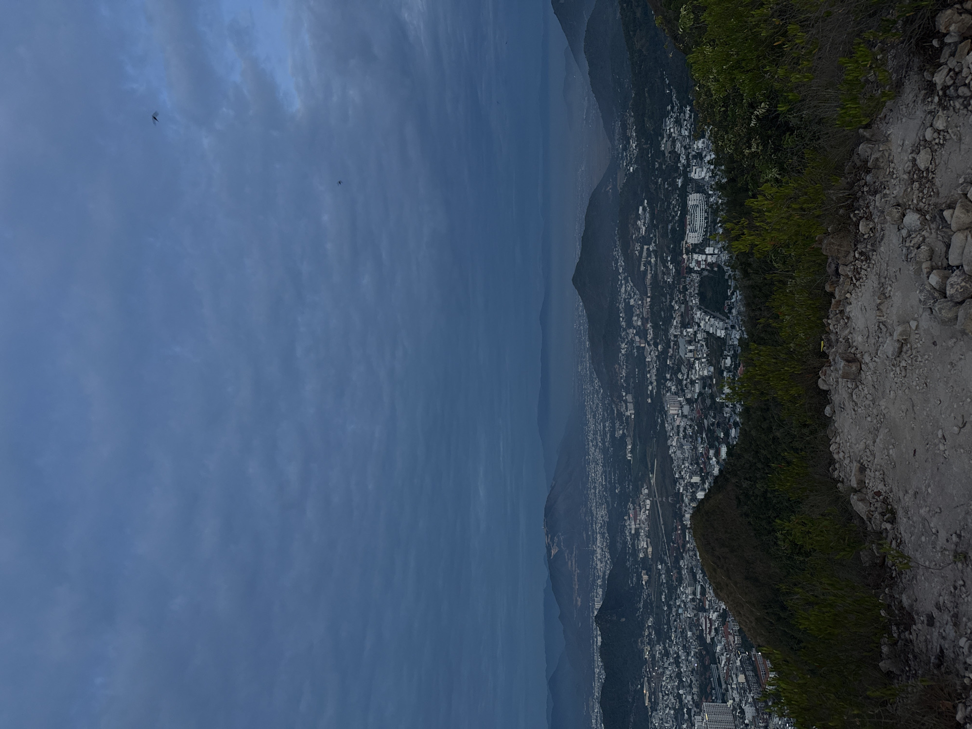 The image is a landscape shot from a high altitude, possibly a mountain or tall building, overlooking a city. The sky is cloudy and overcast, obscuring the sun. Two small objects, possibly birds or insects, are visible in the sky. In the middle ground, a series of mountains leads down to a densely populated city with numerous buildings and streets. On the right side of the image, a rocky, vegetated slope is visible, suggesting the edge of the mountain or high point from which the photo was taken. Bac Nha Trang Ward, Vietnam is likely the location, given the urban density and mountainous terrain. There are no people visible in the image. The overall environment suggests a cloudy day in a mountainous, urban setting.