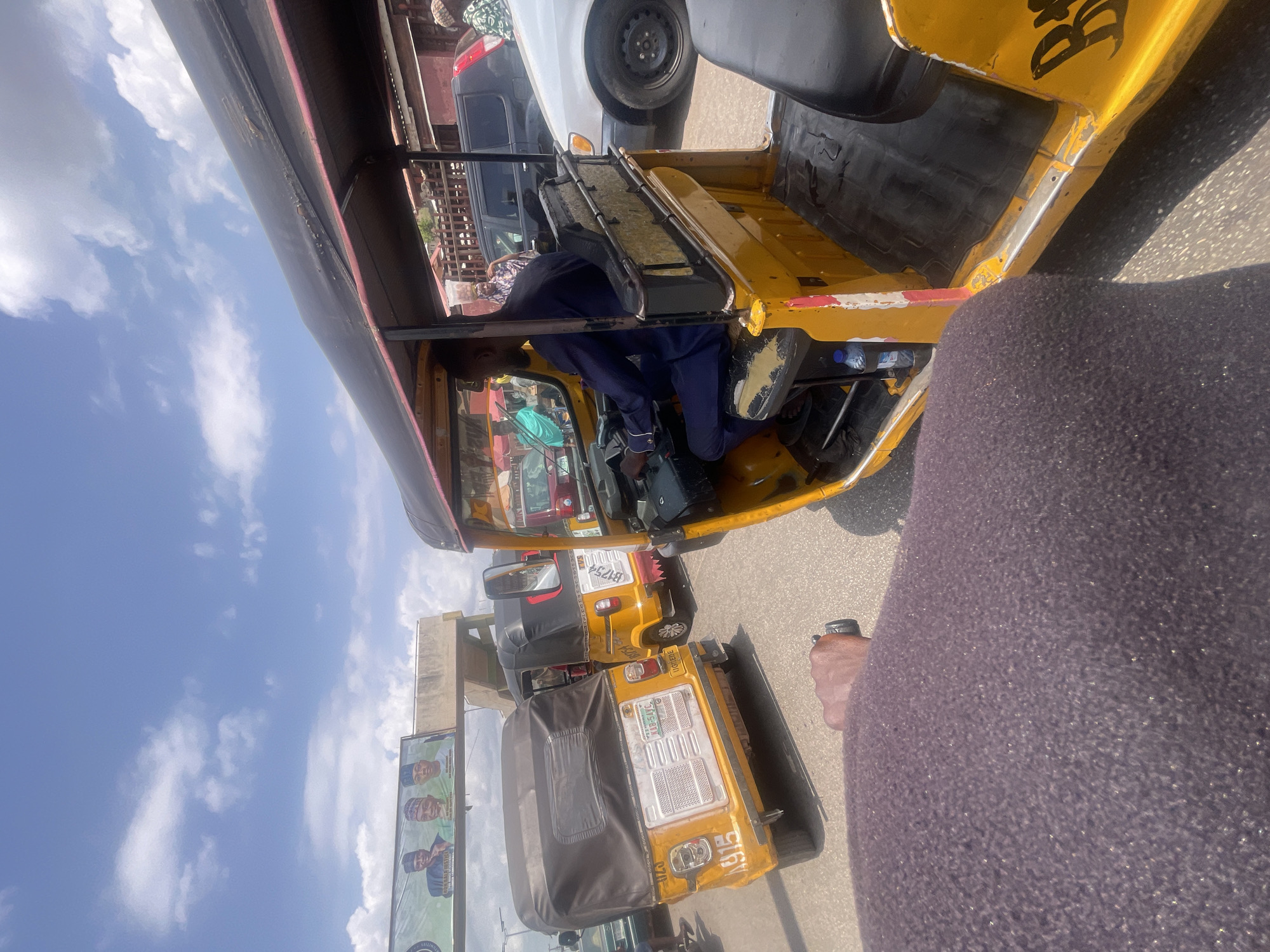 The image, taken at street level, shows a row of yellow auto-rickshaws, commonly known as "Keke Napep," in Lokoja, Nigeria. One rickshaw is partially visible in the foreground, with a person in dark blue clothing seated in the driver's seat. Additional rickshaws are visible behind, aligned along the roadside. A billboard showing several men's portraits is on the left side. The sky is bright blue with scattered clouds. The foreground also contains a portion of a purple-colored article of clothing. The perspective is from inside a rickshaw.