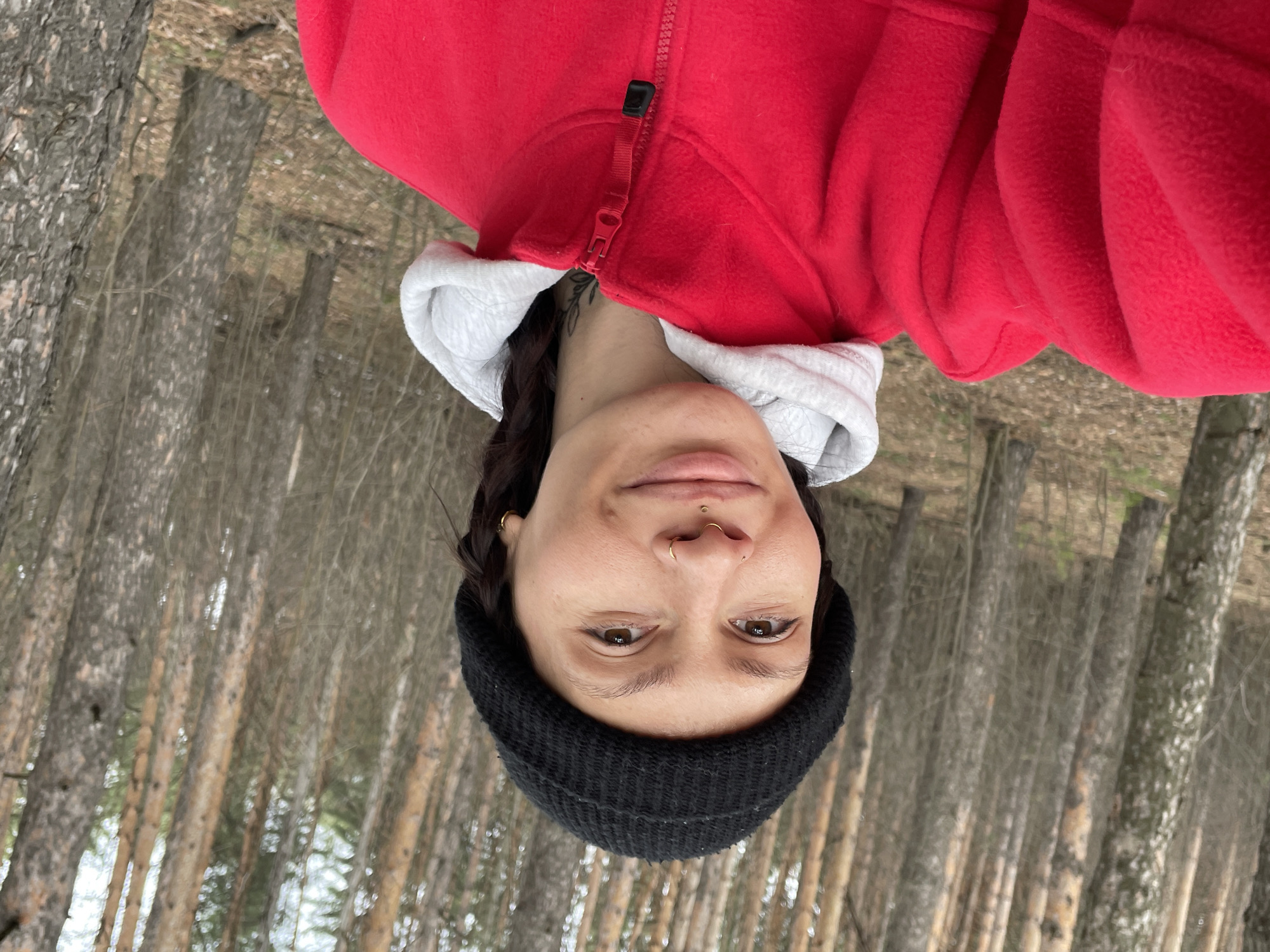A female wearing a black knit hat and a red fleece jacket with a white hood stands in a forest. Tall, slender trees with light bark dominate the background. The scene is potentially located in a forest near Izhevsk, Russia, given the prompt's mention of the city. The female is facing the camera with a neutral expression. The image has been rotated.