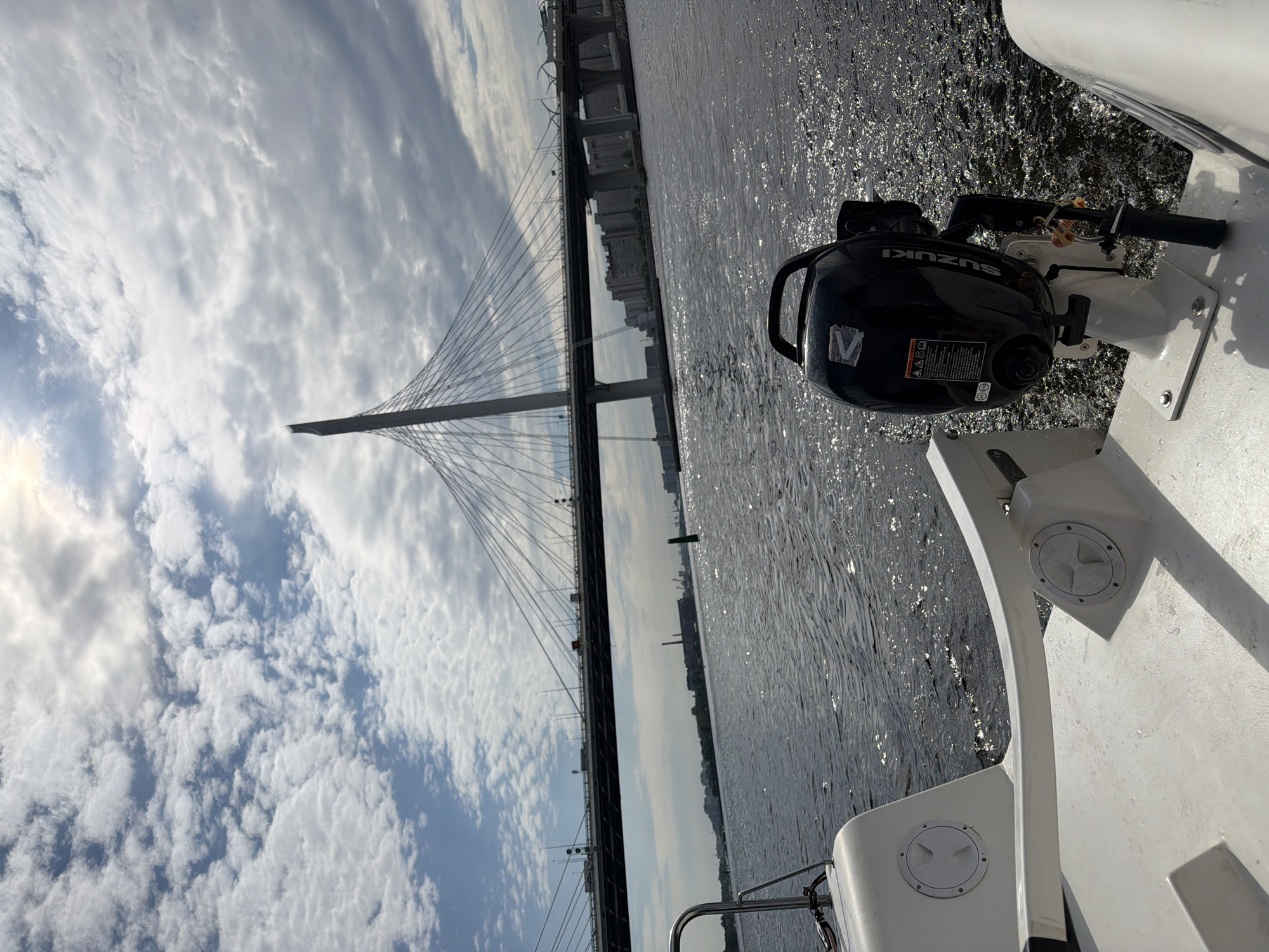 The image shows a view from the stern of a white boat moving on a body of water, likely a river or bay. In the foreground, the rear of the boat is visible with a black outboard motor, identified by the "SUZUKI" logo. The motor has a safety lanyard attached. The water is choppy with reflections of the sky. In the background, a large, modern cable-stayed bridge dominates the horizon, stretching across the water. This bridge is part of the Western High-Speed Diameter in Saint Petersburg, Russia. The sky is cloudy with patches of blue sky and sunlight filtering through. Along the shoreline in the distance, urban buildings and industrial structures can be seen. The scene suggests a daytime excursion on the water with a prominent landmark of the city.