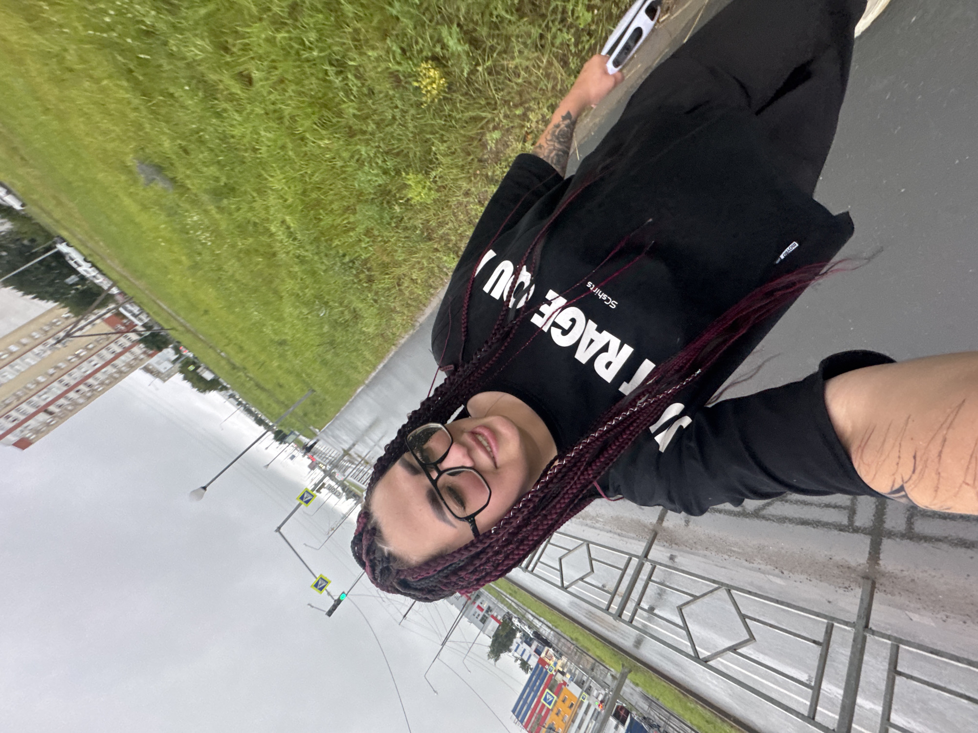 A young woman with dark, braided hair and glasses is taking a selfie outdoors on a cloudy day. She is wearing a black t-shirt with white text that reads "COURAGE YOU." She has tattoos on her left arm. She is holding a white and black object in her right hand. The background shows a wet sidewalk, a metal fence, a grassy area, streetlights, traffic signals, and buildings, suggesting an urban environment. The scene appears to be a street in Tolyatti, Russia, possibly near a residential area with a commercial building in the distance. The wet ground indicates recent rain.