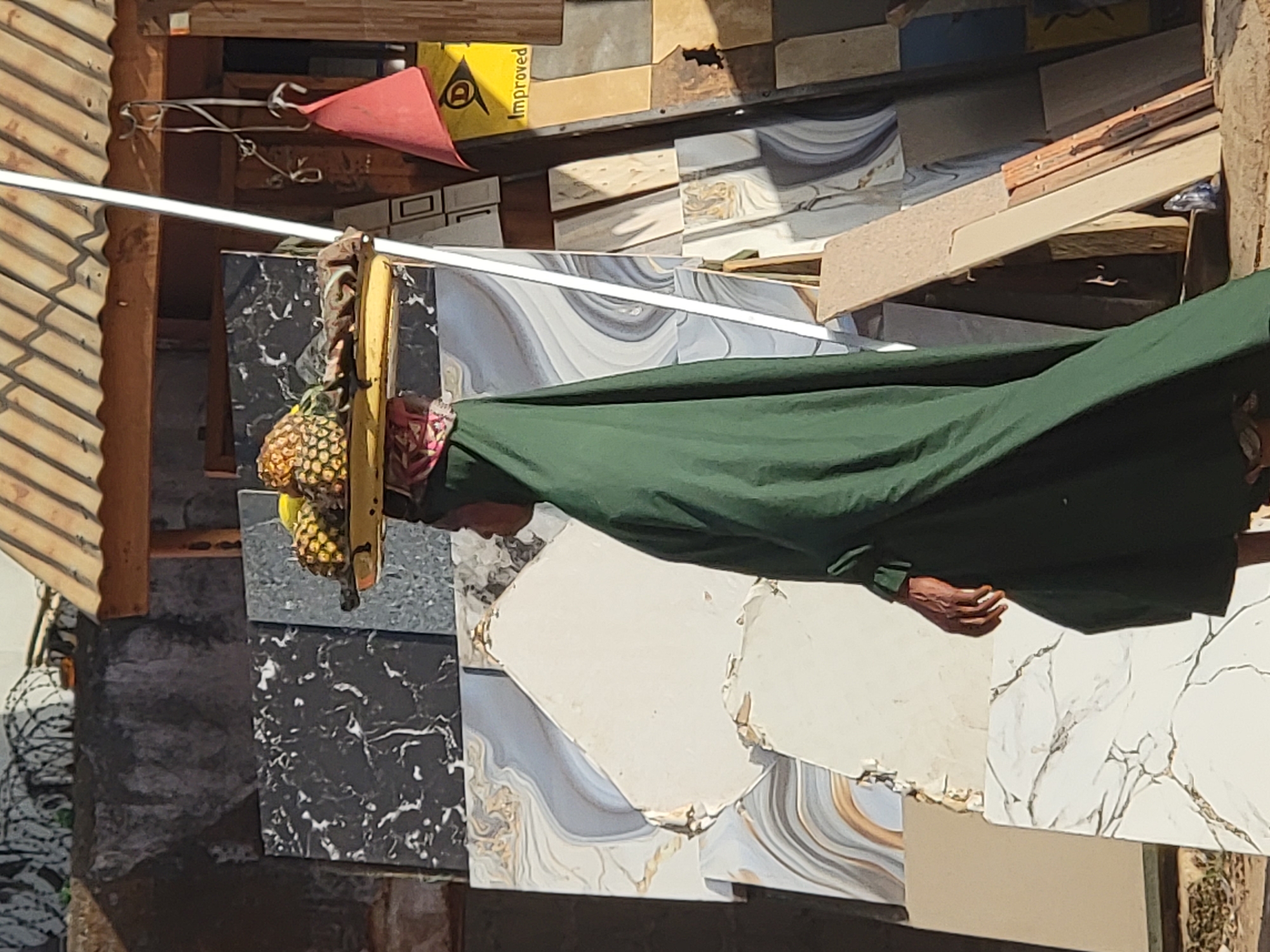 In Maiburiji, Nigeria, the image captures an individual wearing a green garment. The person is positioned near a display of various tiles, some patterned in black marble and others with swirling grey and gold designs. A rack of pineapples hangs nearby. The environment consists of a mix of building materials and finished tiles, suggesting a retail or construction-related location. The scene is captured in daylight, casting shadows across the subjects.