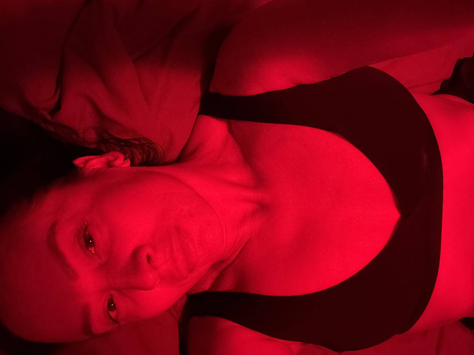 A woman lies in bed wearing a black bra. The scene is lit with a strong red light, which obscures details. No information suggests the location is Chaikovsky, Russia.