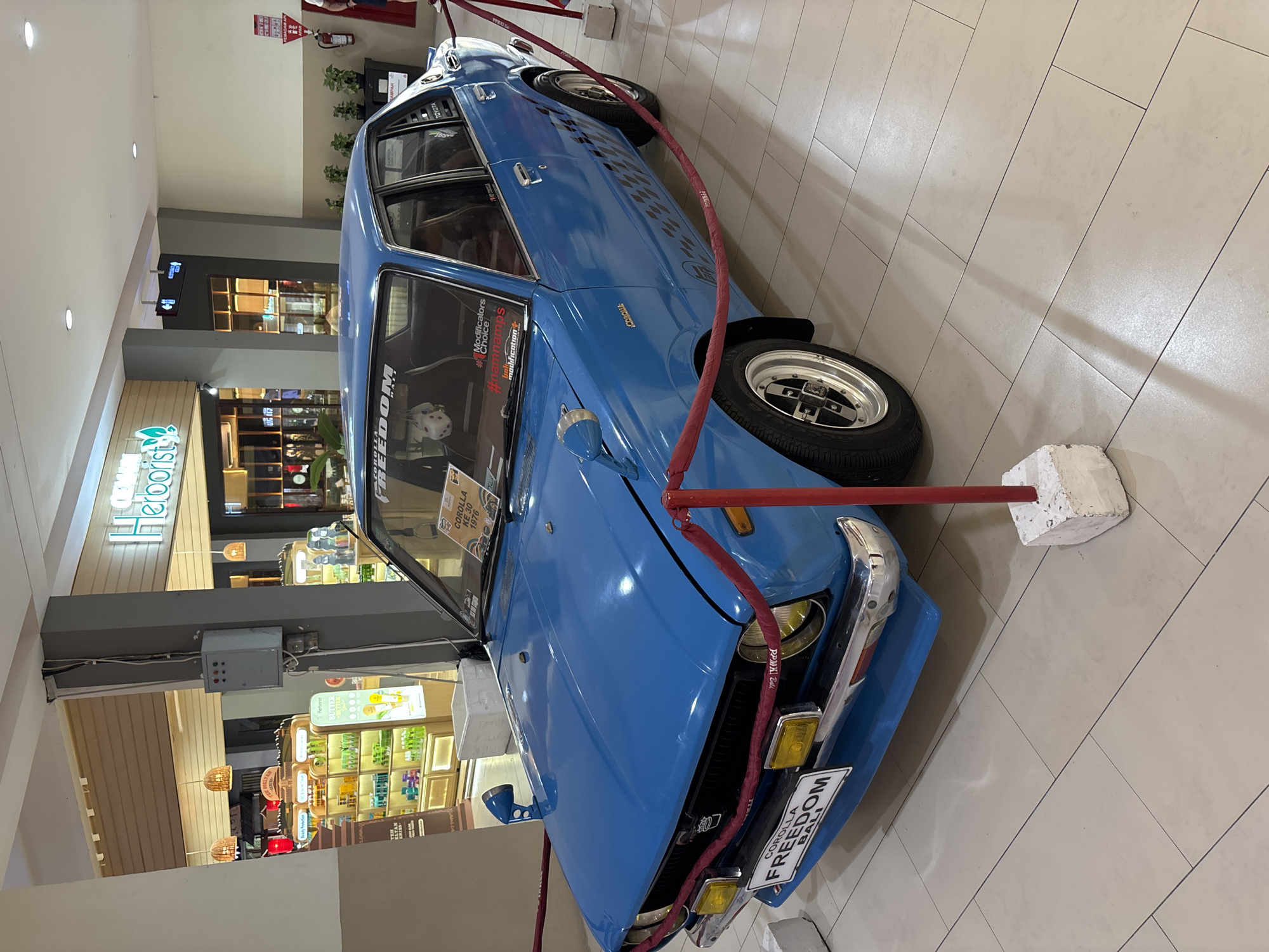 The image shows a blue 1976 Toyota Corolla KE30 displayed in an indoor shopping mall setting in Kuta, Bali, Indonesia. The car is surrounded by red rope barriers secured with concrete blocks. A sign in the windshield provides information about the vehicle. The car's license plate reads "COROLLA FREEDOM BALI." Several retail storefronts with products on display are visible in the background.