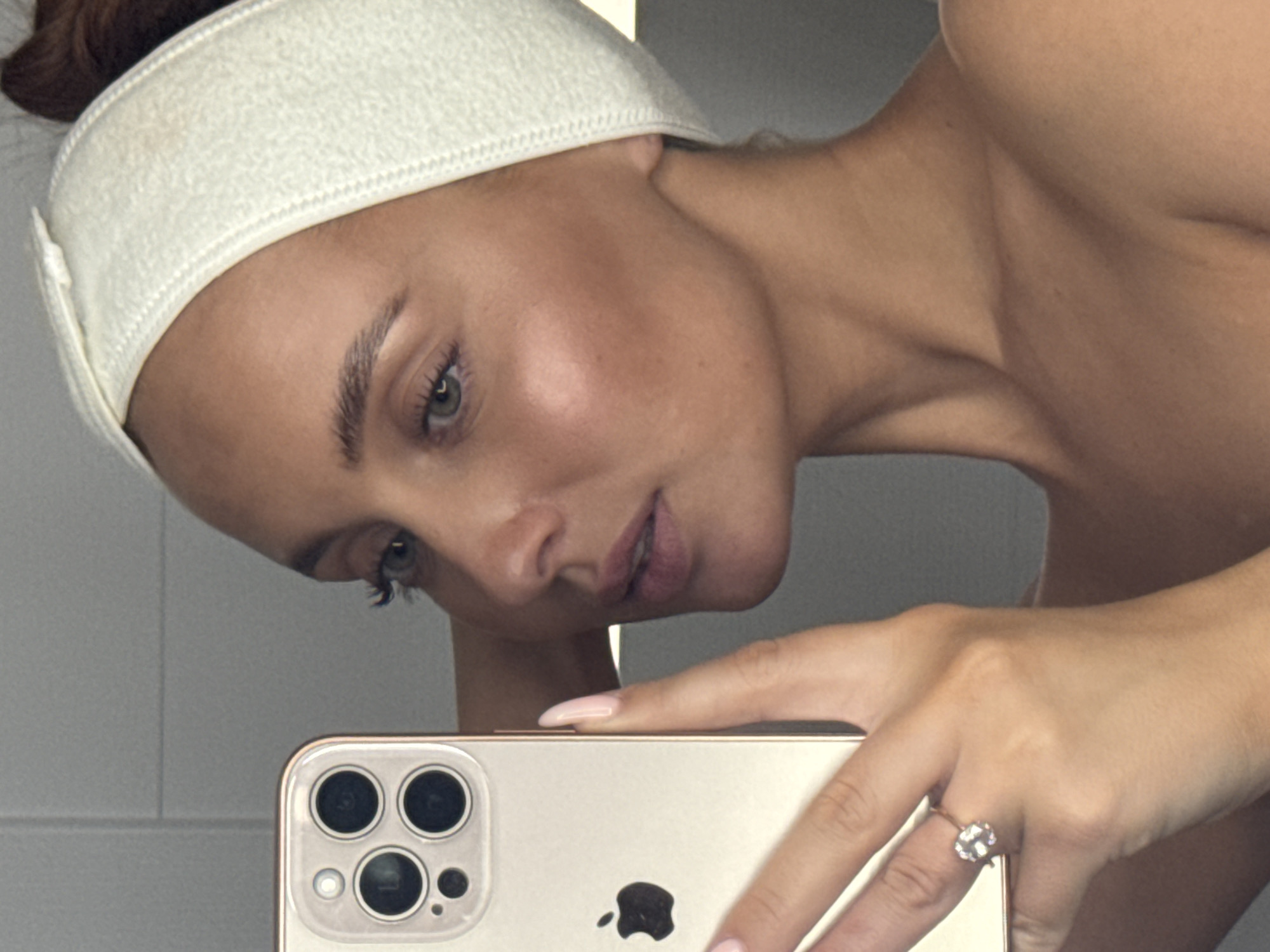 The image shows a close-up, selfie-style view of a woman's face and upper body. She is wearing a light-colored fabric headband and has her hair pulled back. Her skin appears to have a tanned complexion with visible contouring makeup on her cheeks. She is holding a gold iPhone in her left hand, which is reflected in a mirror, obscuring part of her face. A diamond ring is visible on her left ring finger. The background is a gray, tiled wall, suggesting an indoor setting such as a bathroom. There is no visible indication of the city of Chaikovsky, Russia.