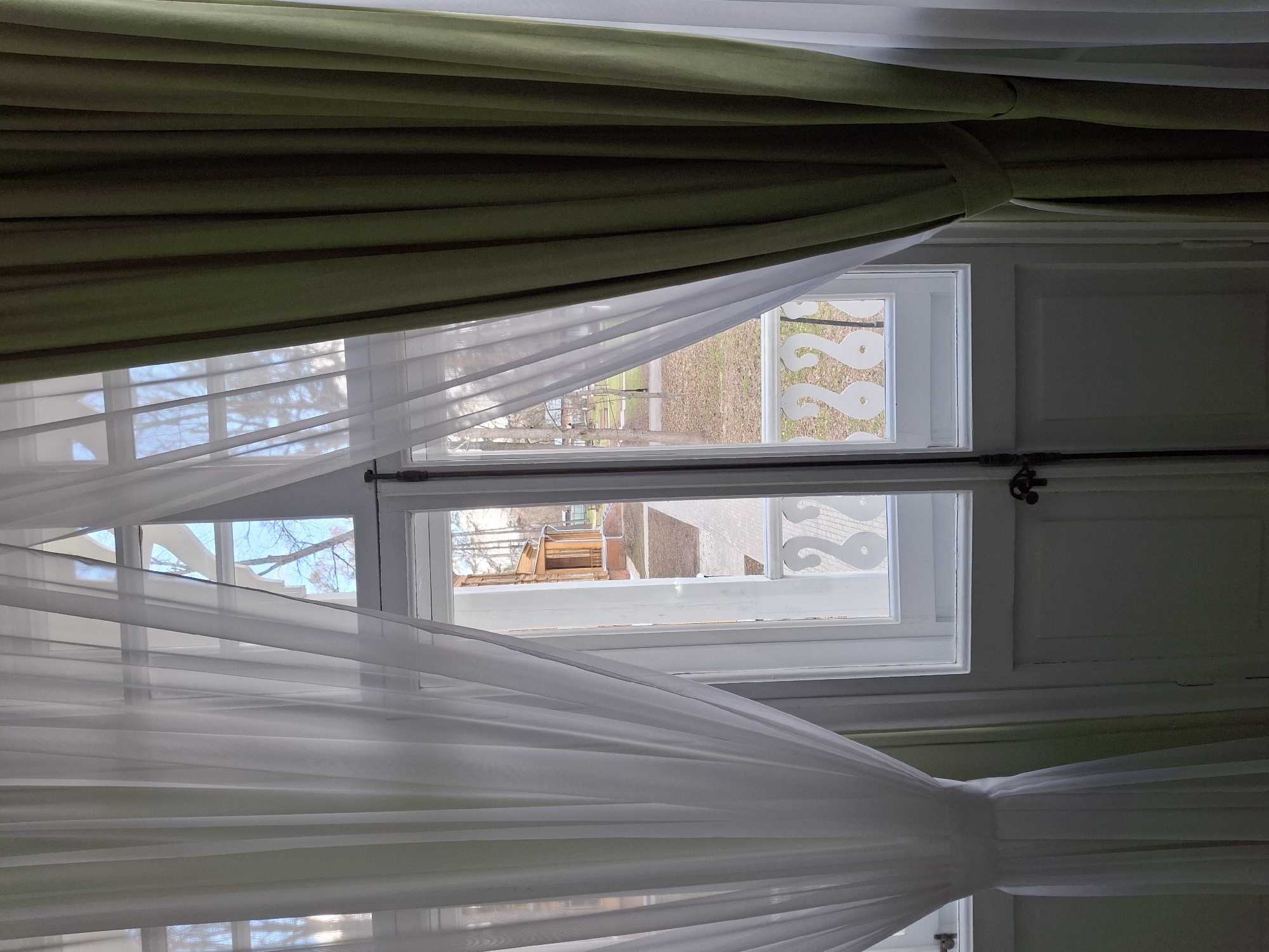 The view from inside a building in Borok, Russia, features a double-hung window partially obscured by olive-green and sheer white curtains. The upper window reveals a grassy yard with leafless trees and a glimpse of neighboring buildings, one with a decorative trim. The lower window offers a similar view, highlighting a rooftop and a building in the background. A decorative white trim with a swirl pattern is visible on one side of the upper window. The overall scene is a quiet, interior view looking out onto an early spring day in a residential area.
